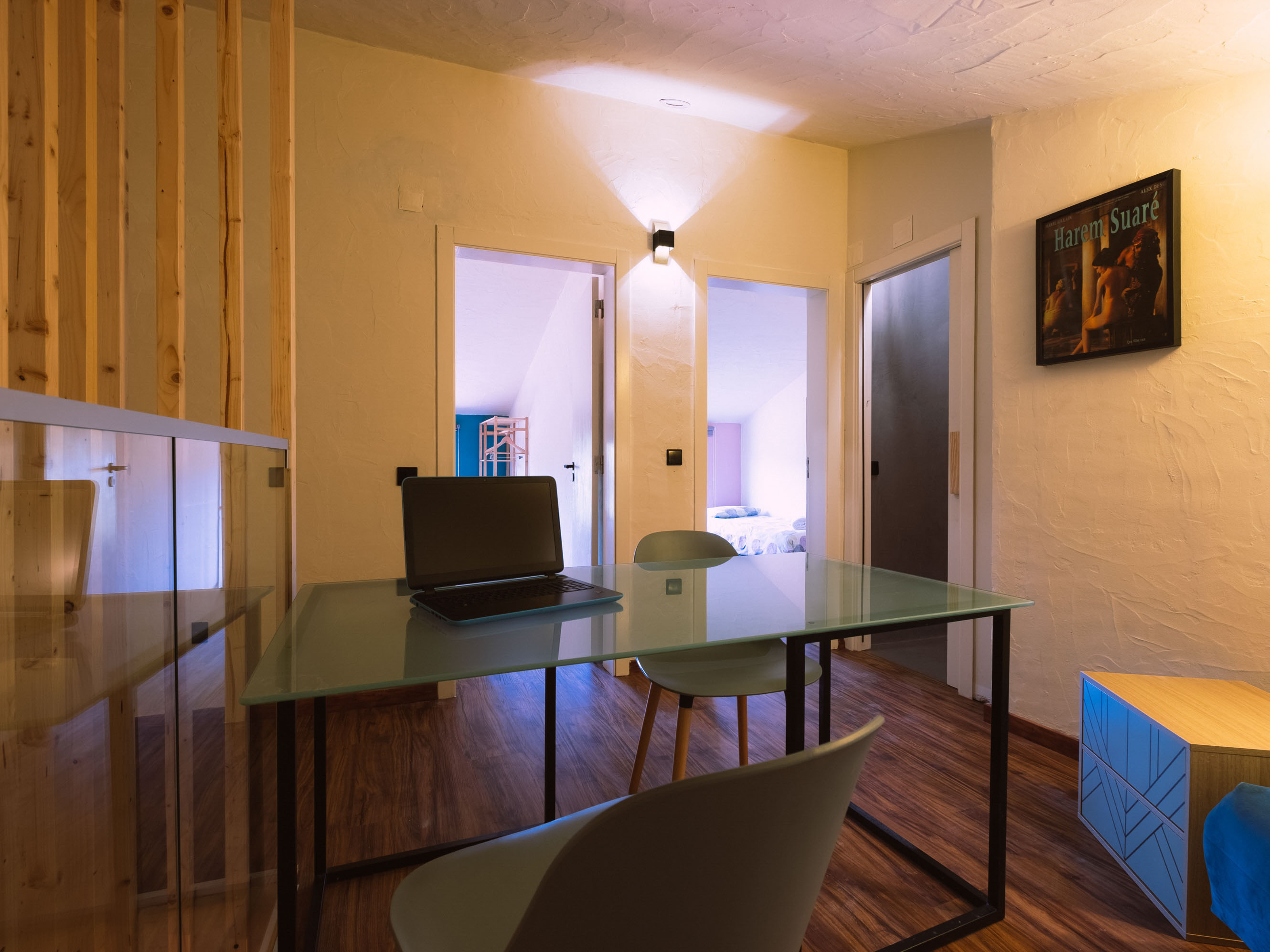 Room with glass table, chairs, laptop, wooden floor, two doorways, and a framed picture on white wall.