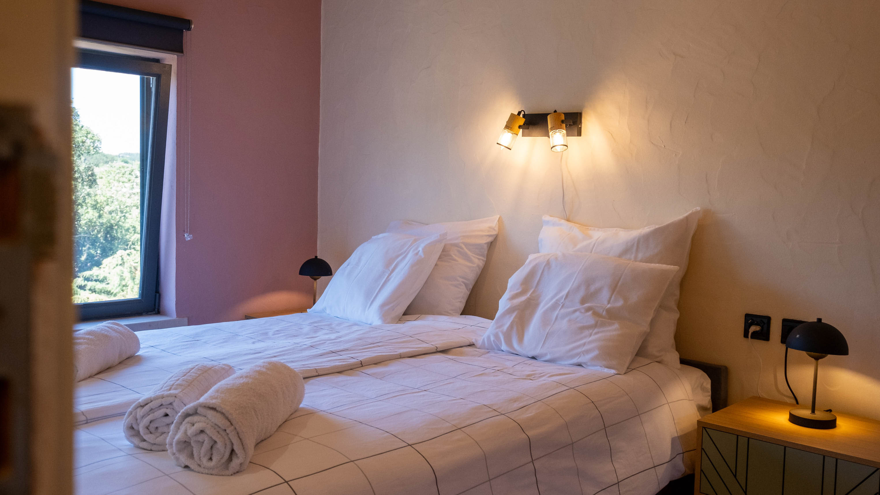 Bed with white linens, pillows, rolled towels, bedside lamps, and a window showing outdoor greenery.