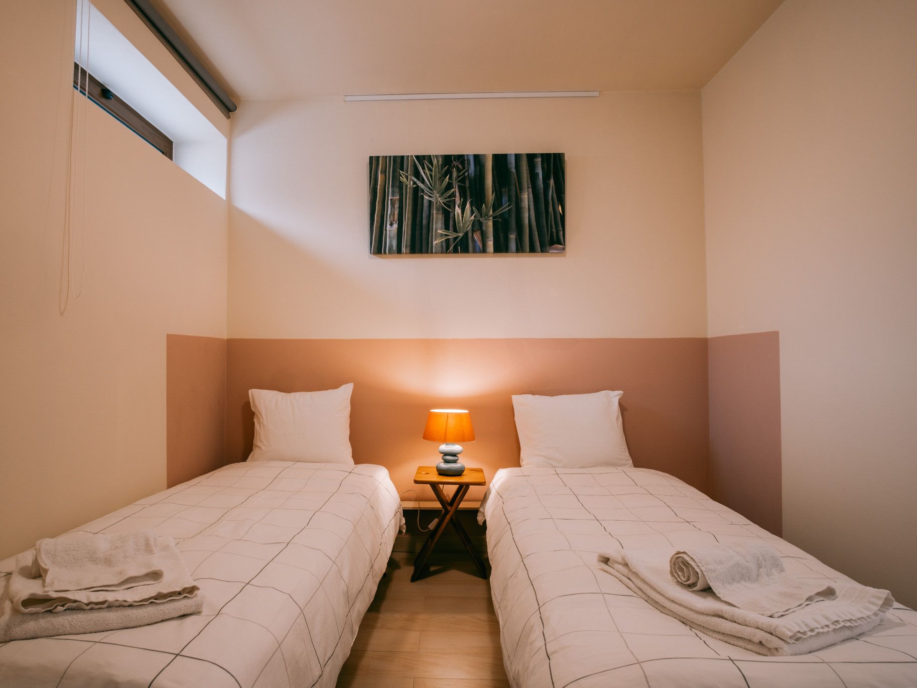 Two single beds with white linens, bedside table lamp, folded towels, bamboo artwork on pale pink wall.