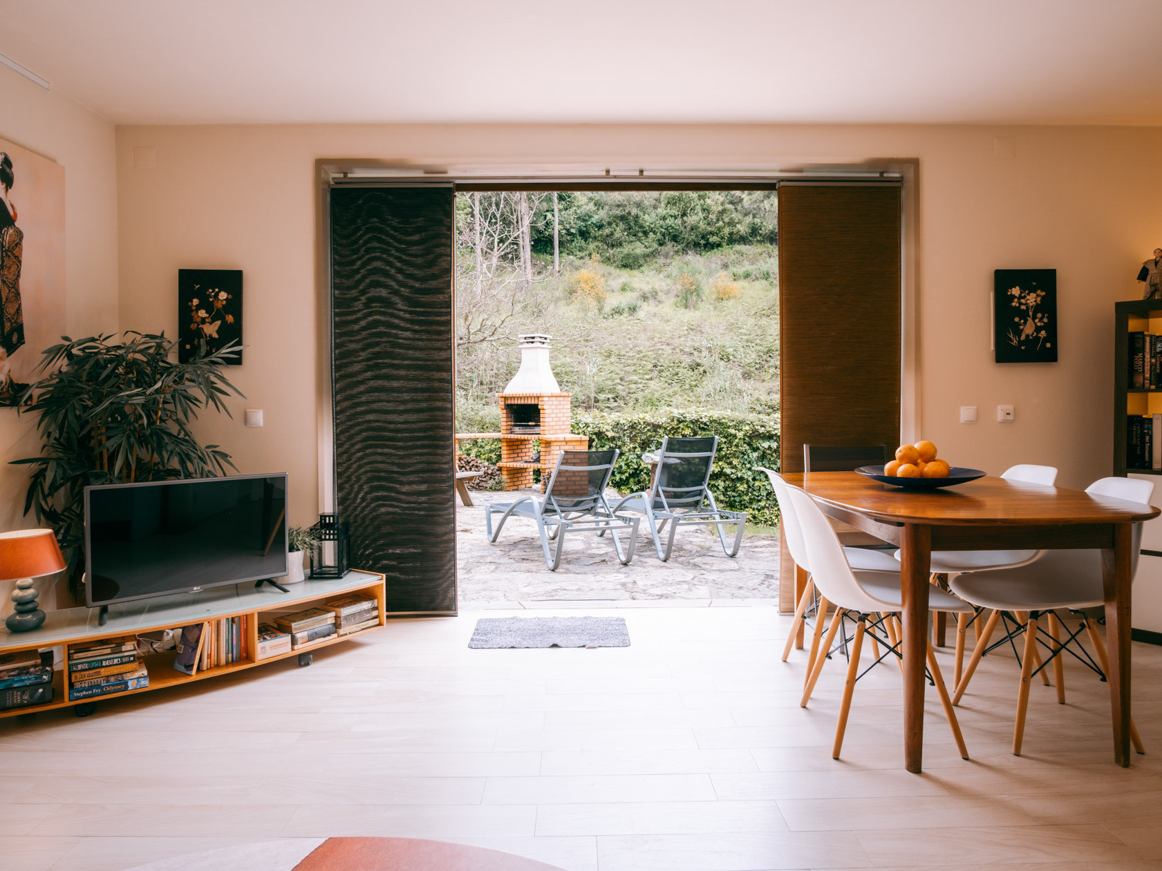 Living-dining room with TV, table and fruit bowl, sliding doors to patio with lounge chairs and brick grill.