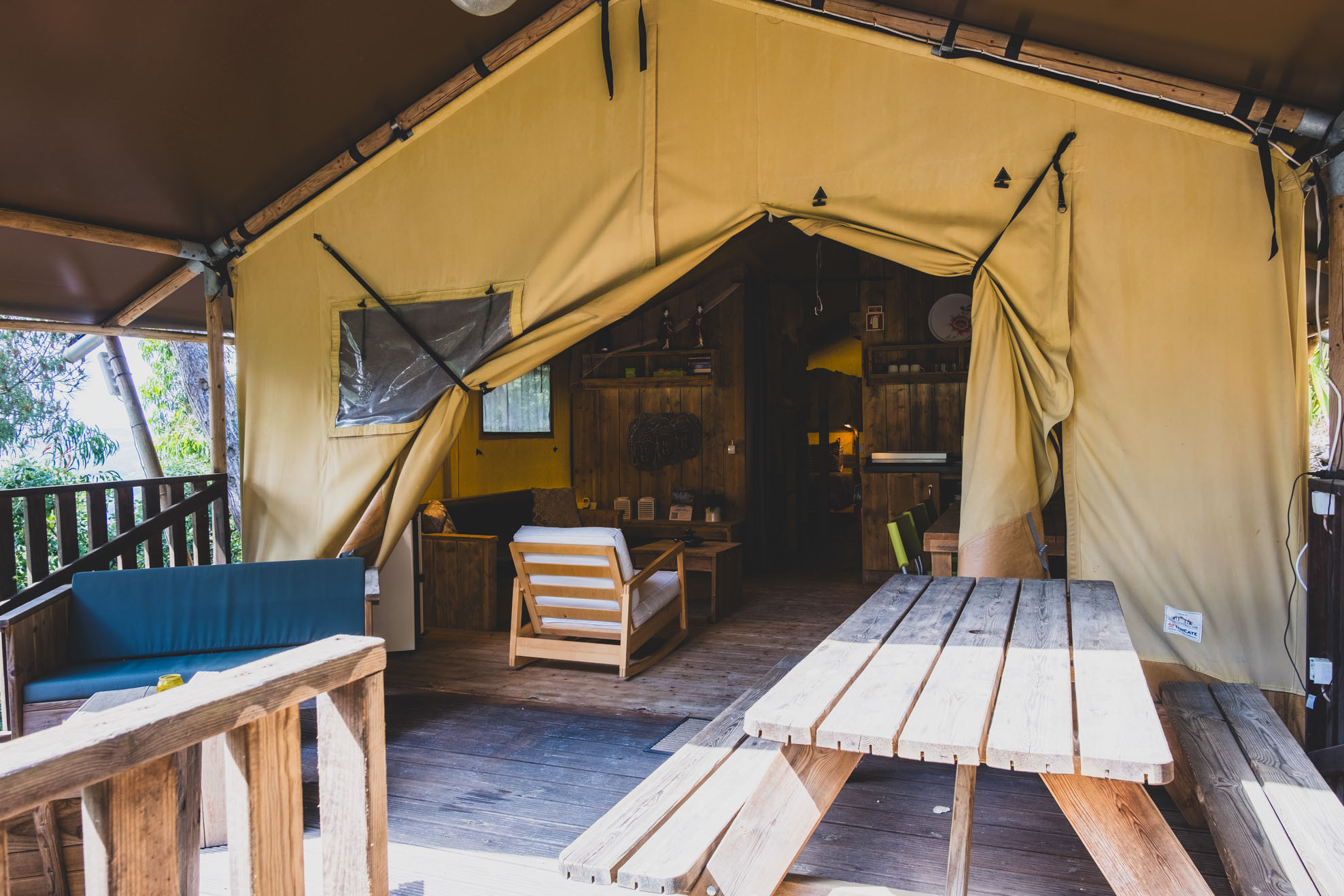 Wooden picnic table and benches outside a large beige tent with a seating area inside.