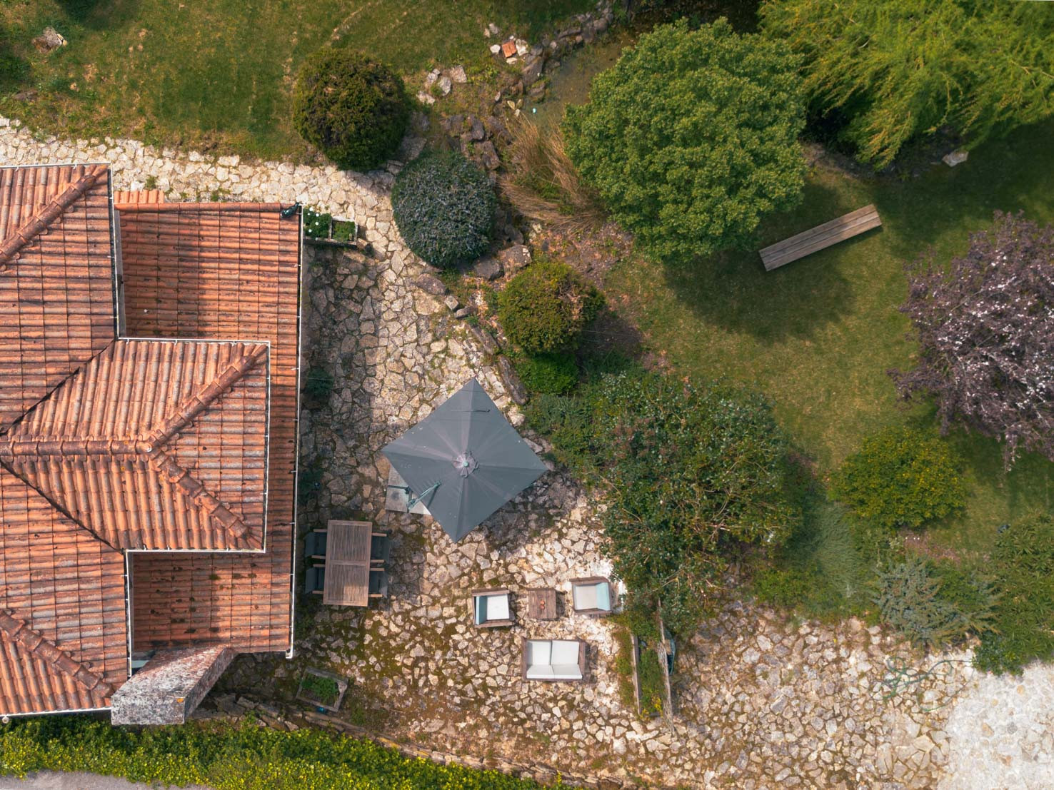 Terracotta-roofed building next to stone patio with umbrella, chairs, tables, and surrounding greenery.
