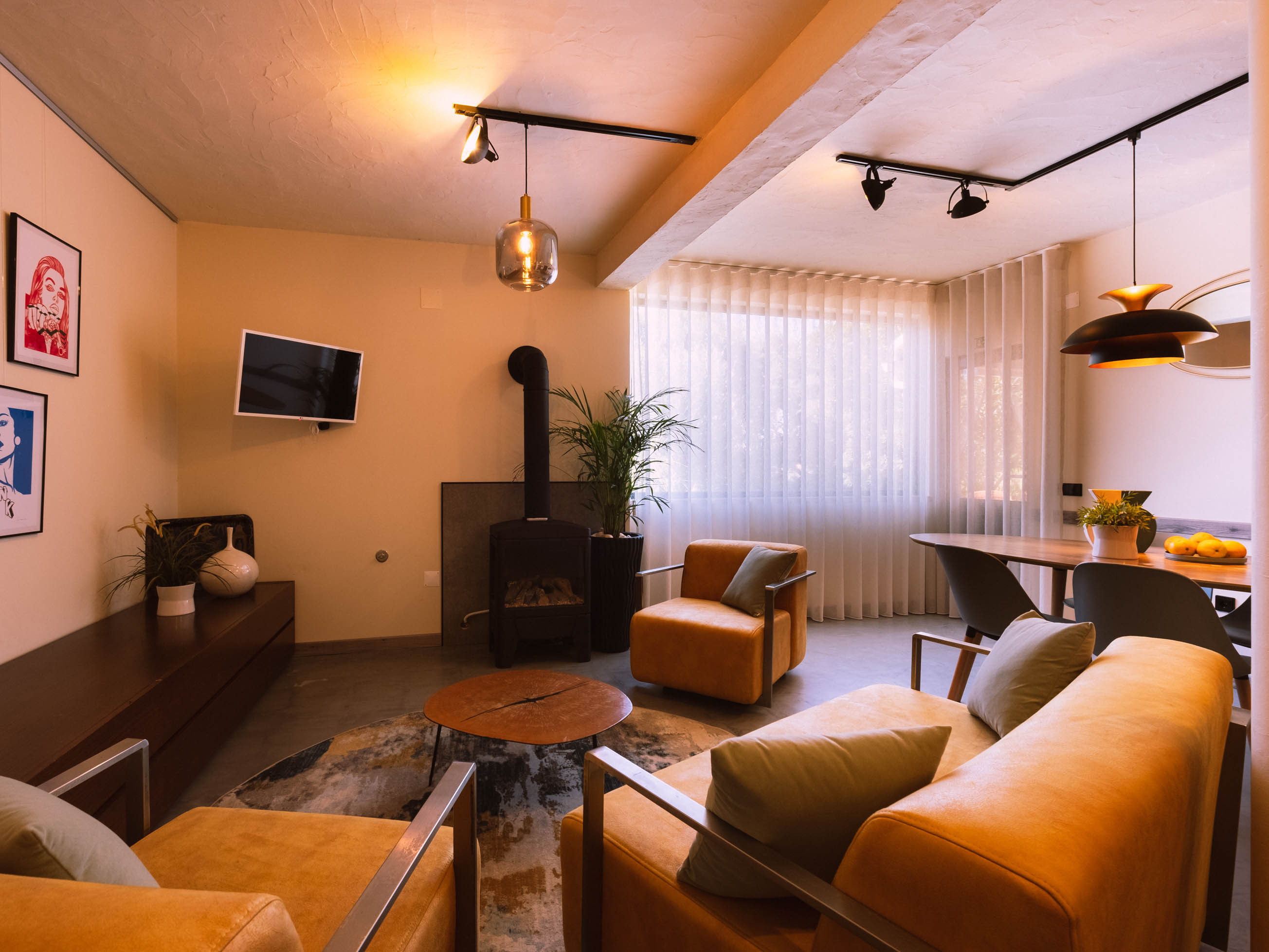 Living room with orange armchairs, sofa, wood stove, round wooden table, TV, dining table, and large window