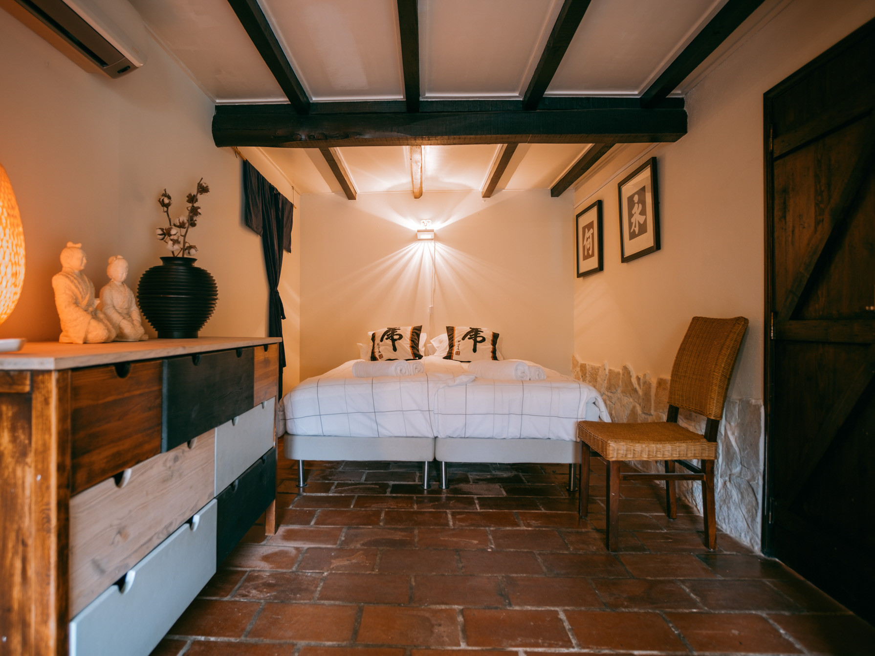 Room with twin beds, wooden beams, tile floor, dresser with decorations, chair, and framed art on walls.