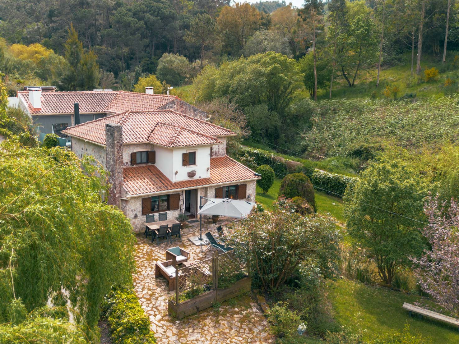 House with terracotta roof tiles surrounded by green trees and a stone-paved patio with outdoor furniture.