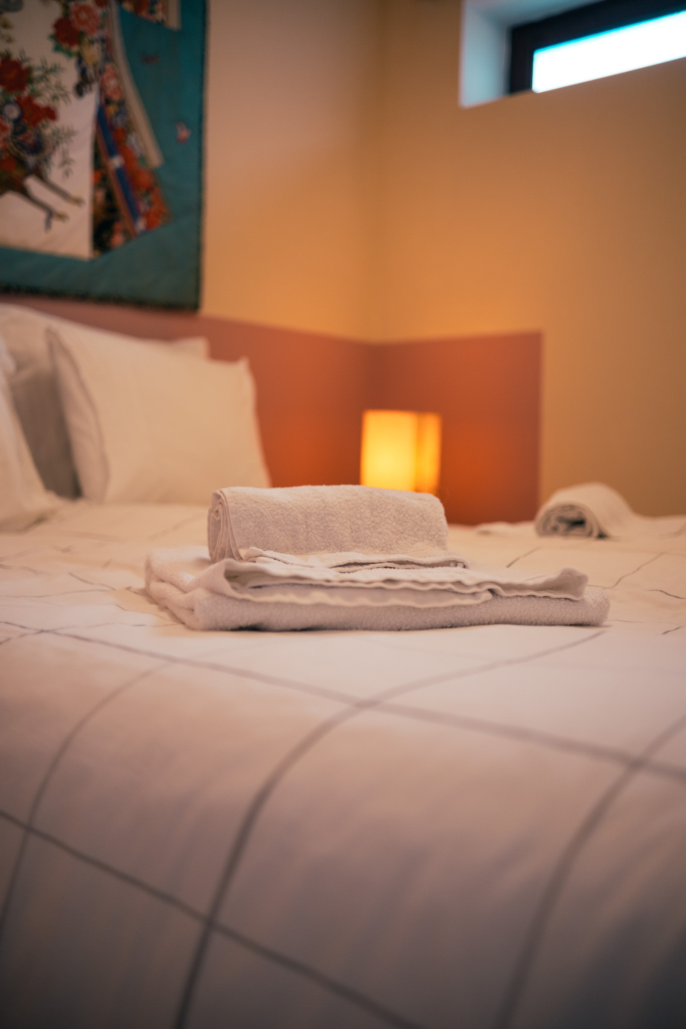 Folded towels placed on a bed with pillows, a glowing bedside lamp, and a wall hanging.