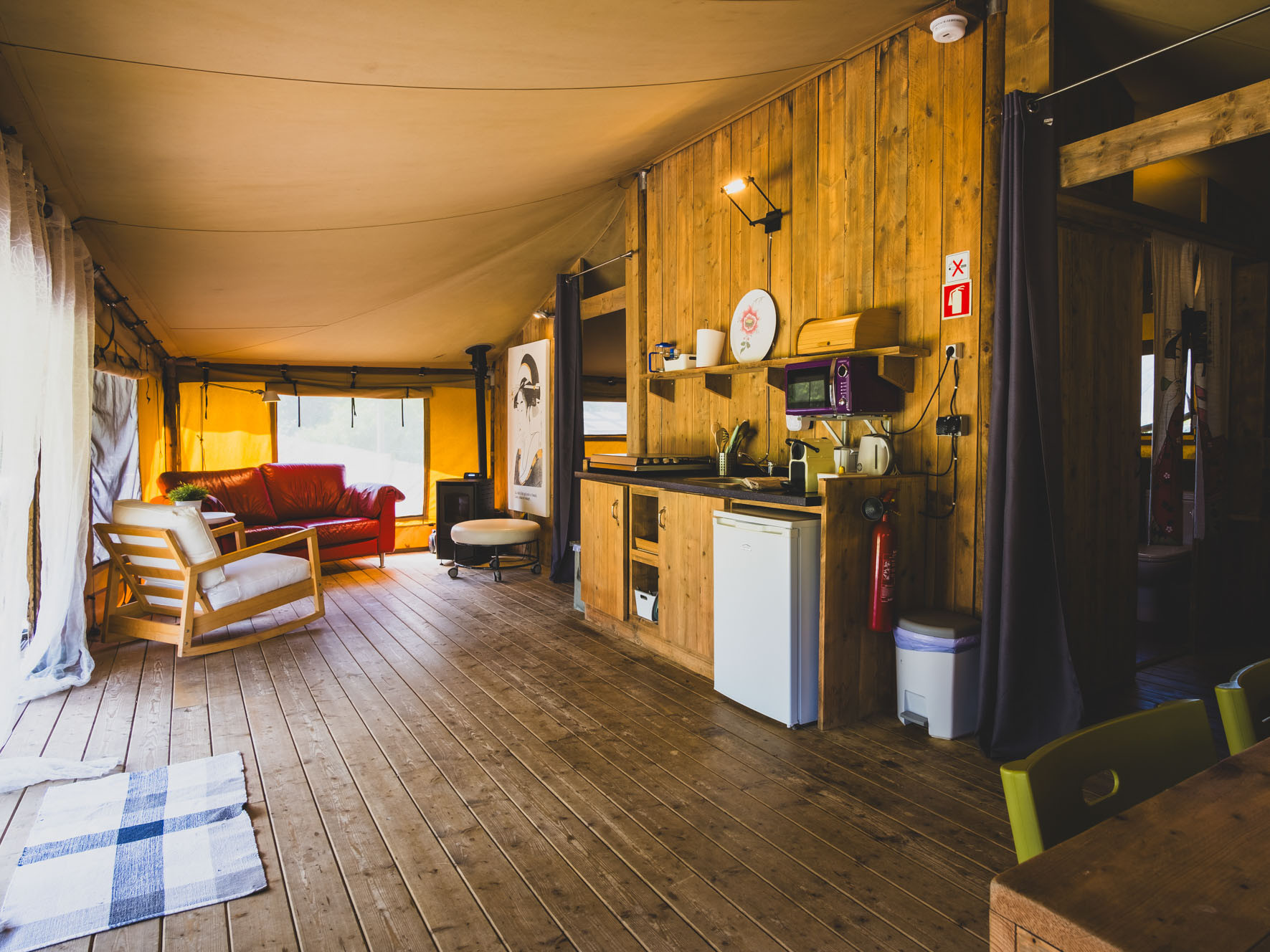 Spacious wooden interior with kitchen area, armchair, sofa, and large windows covered by curtains.