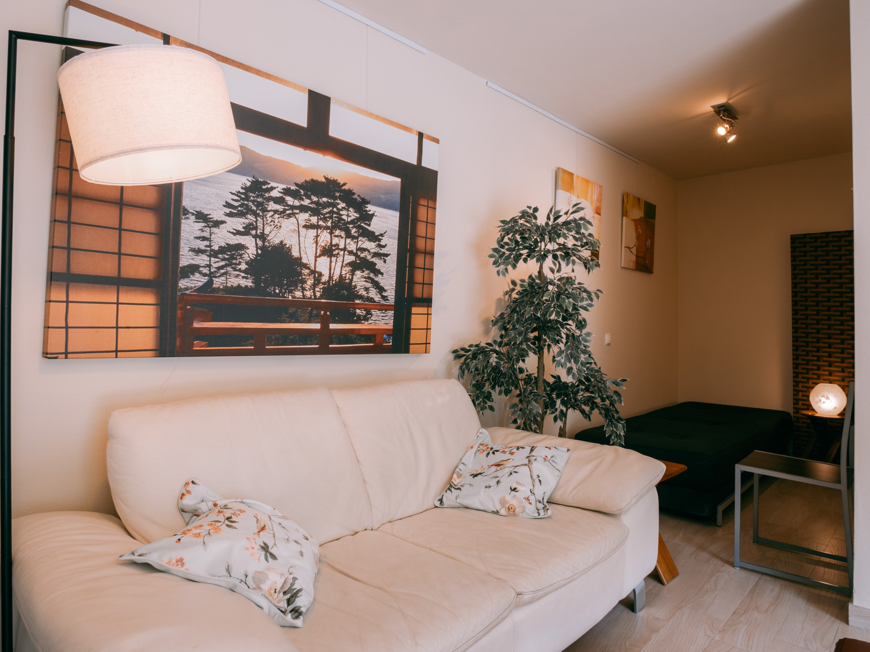 Beige sofa with floral cushions under framed tree landscape, floor lamp, potted plant, side table and dark daybed.