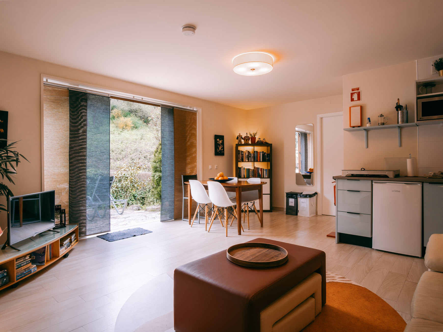 Open-plan living room with dining table, white chairs, kitchenette, TV unit, bookshelf and sliding glass doors to greenery.