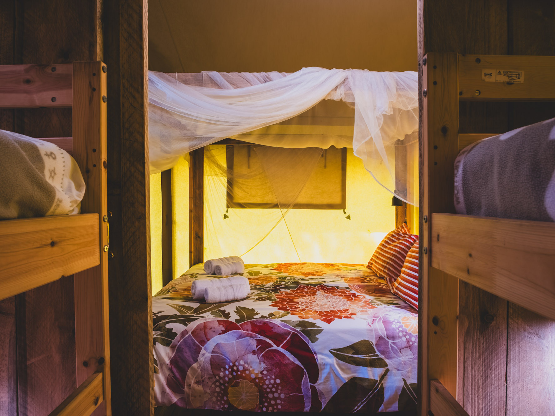 Cozy bed with floral bedding and a white mosquito net between wooden bunk beds in a softly lit