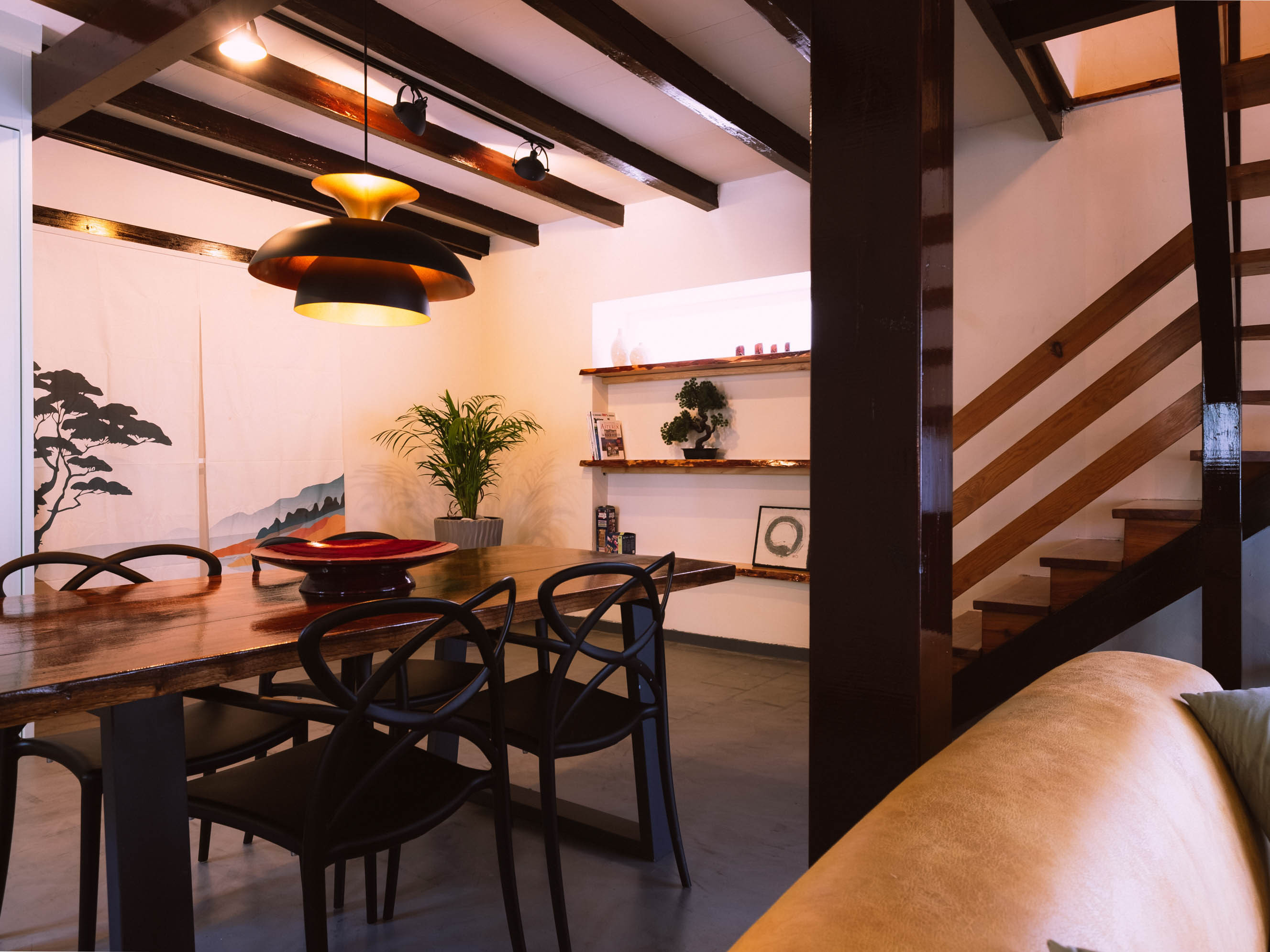 Dining area with wooden table, six black chairs, modern pendant light, staircase, and decorative plant.