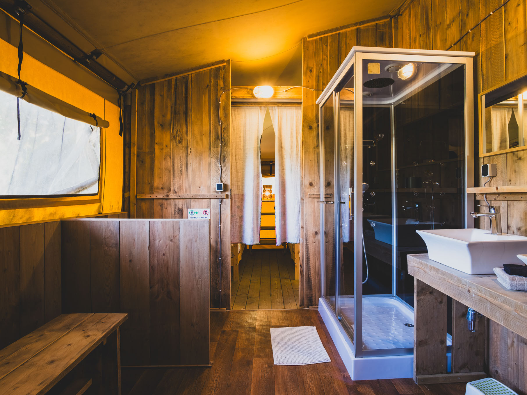 Interior of a wooden cabin bathroom with glass shower, sink, wooden bench, and fabric window covering.