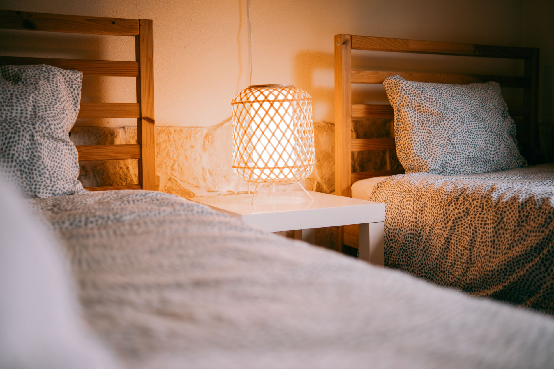 Two single beds with patterned bedding separated by a small white nightstand with a lit lamp.