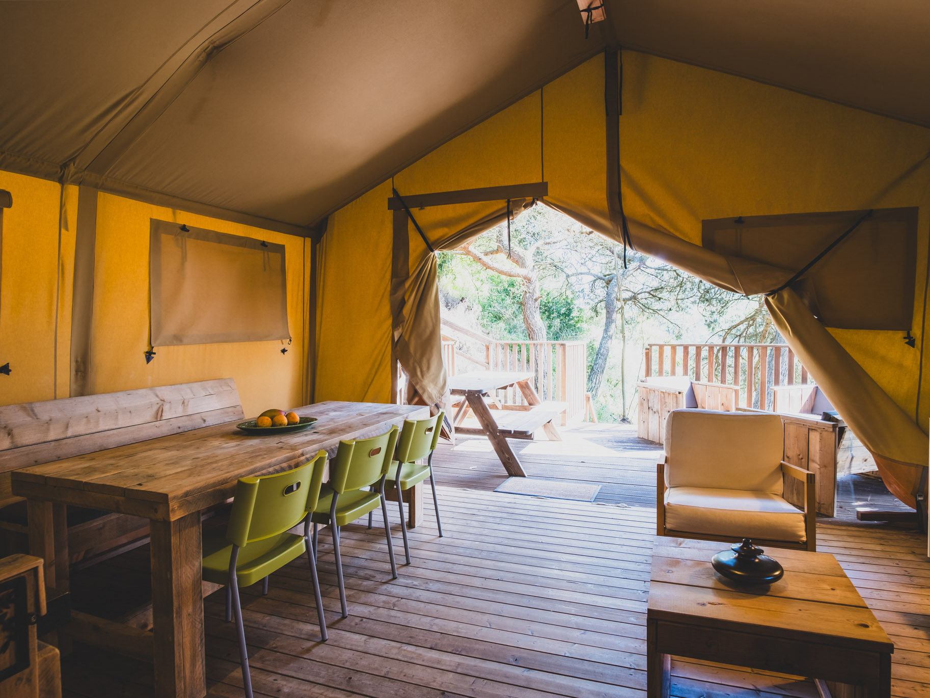 Interior of a large yellow tent with wooden dining table, green chairs, armchair, and wooden deck outside.