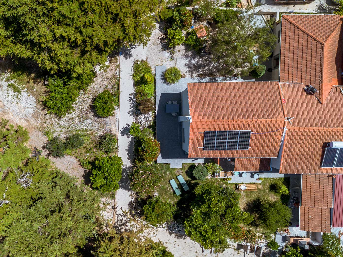 Top-down view of a house with a red tiled roof and solar panels surrounded by trees and garden