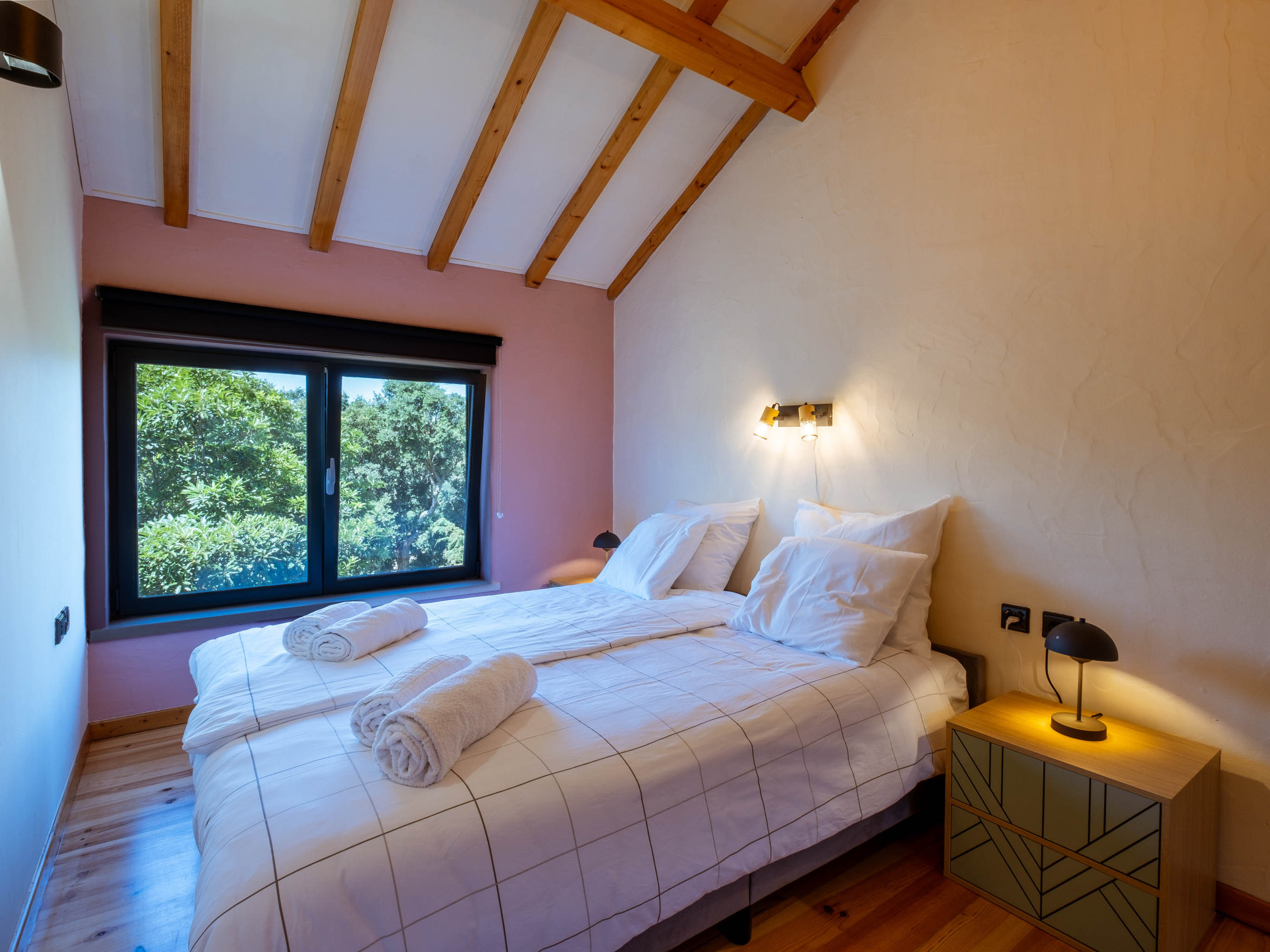 Cozy bedroom with two single beds, white checkered bedding, rolled towels, wooden ceiling beams, window with green outdoor