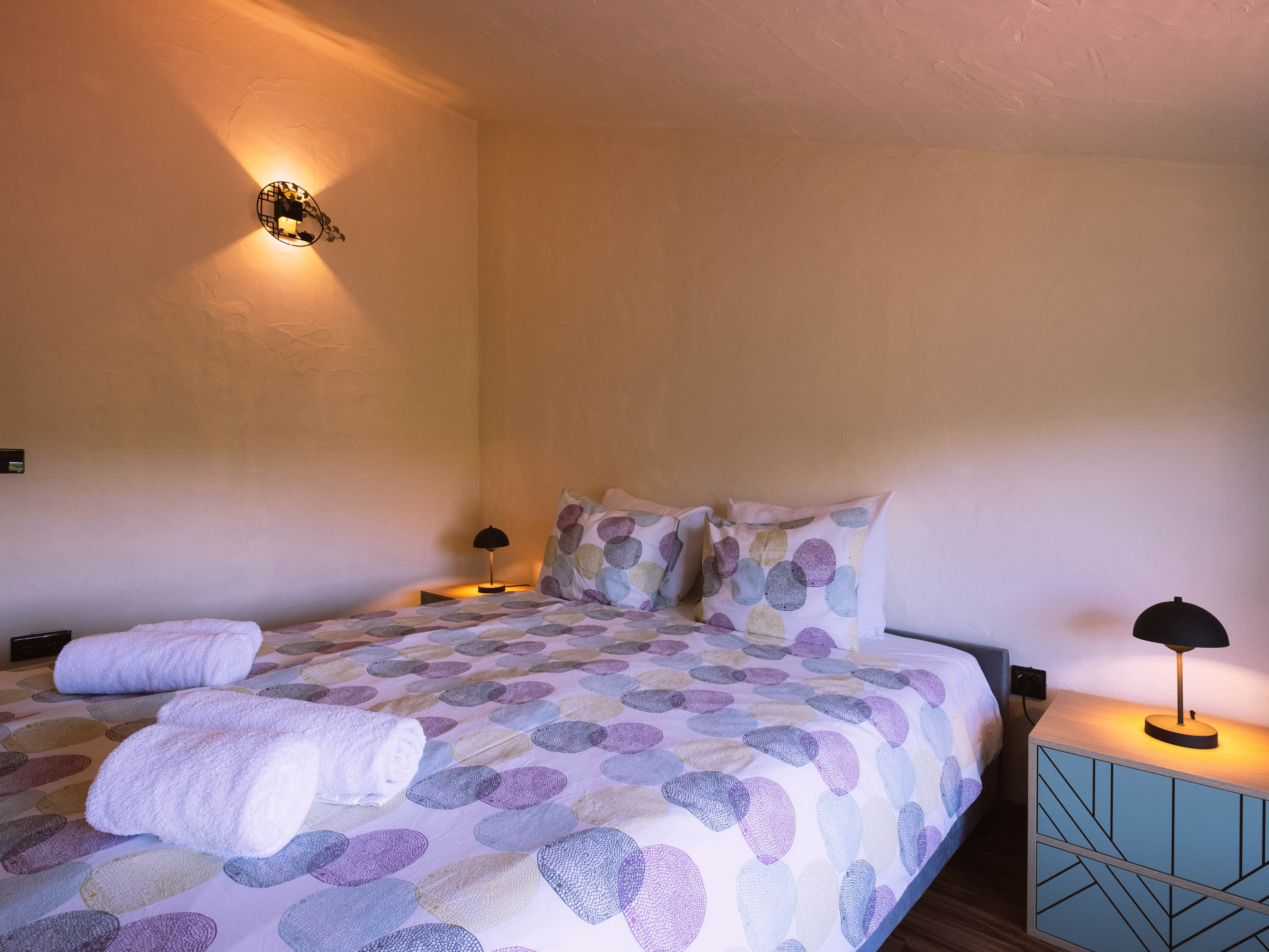 Bed with pastel-colored circle patterned bedding, two rolled white towels, small bedside lamps, and a wall light.
