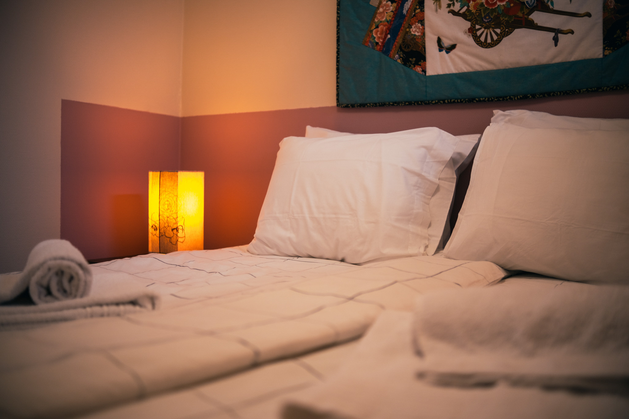 Bed with white pillows and folded towels beside a lit bedside lamp and wall hanging.