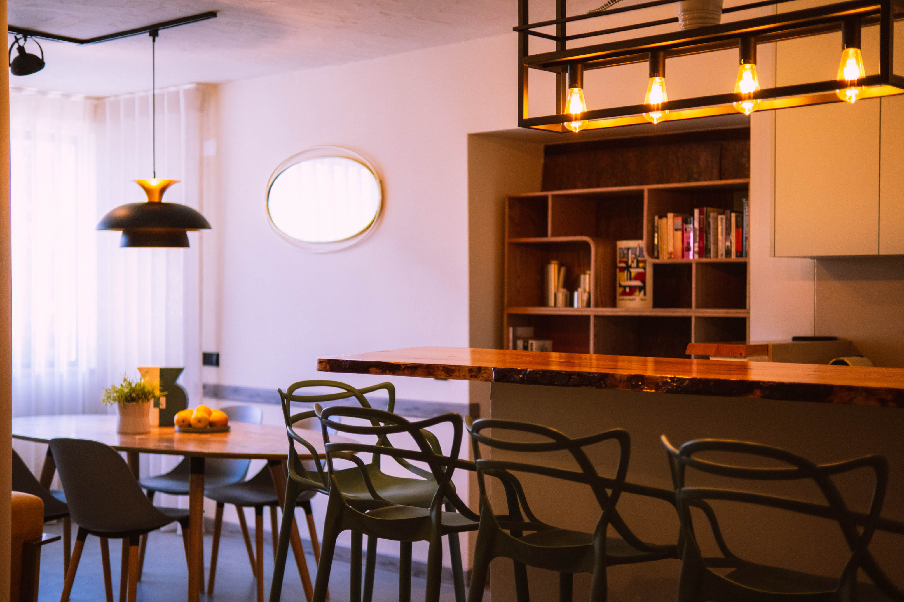 Modern dining area with wooden table, multiple chairs, pendant lights, and a bookshelf with books.