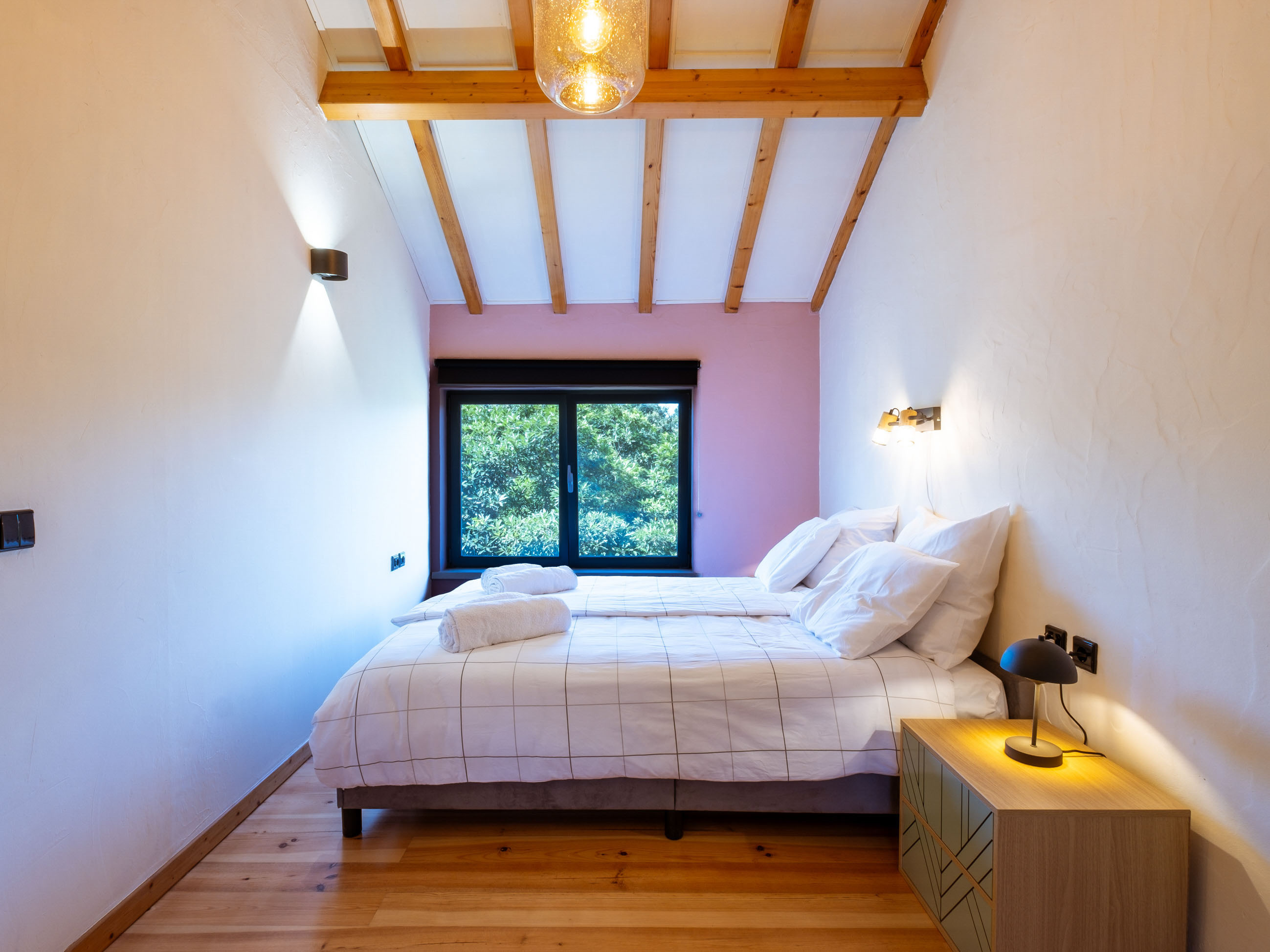 Bedroom with two single beds, wooden floor, large window with green view, and wooden ceiling beams.