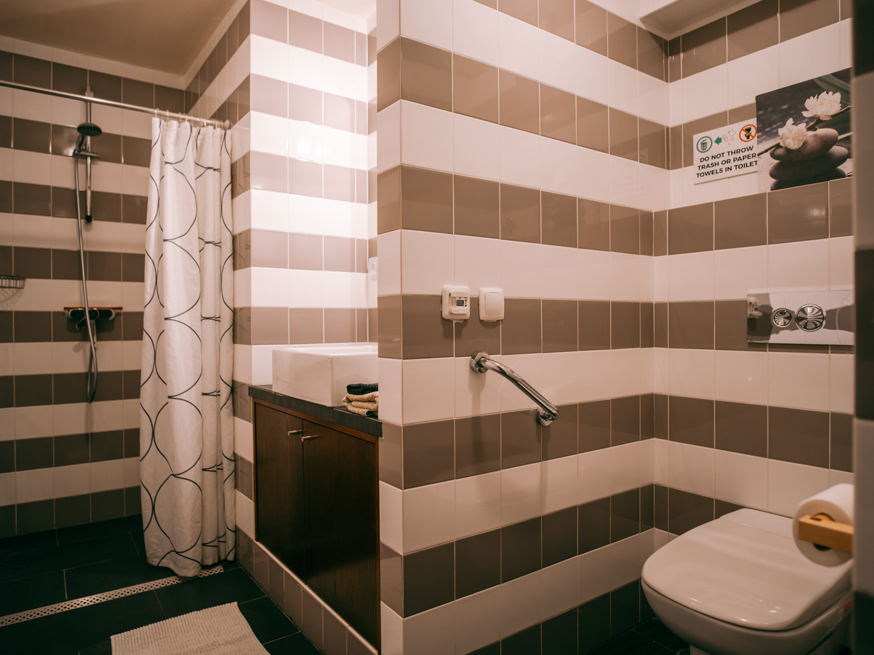 Bathroom with striped beige and white tiles, shower with curtain, sink, and toilet with support rail.