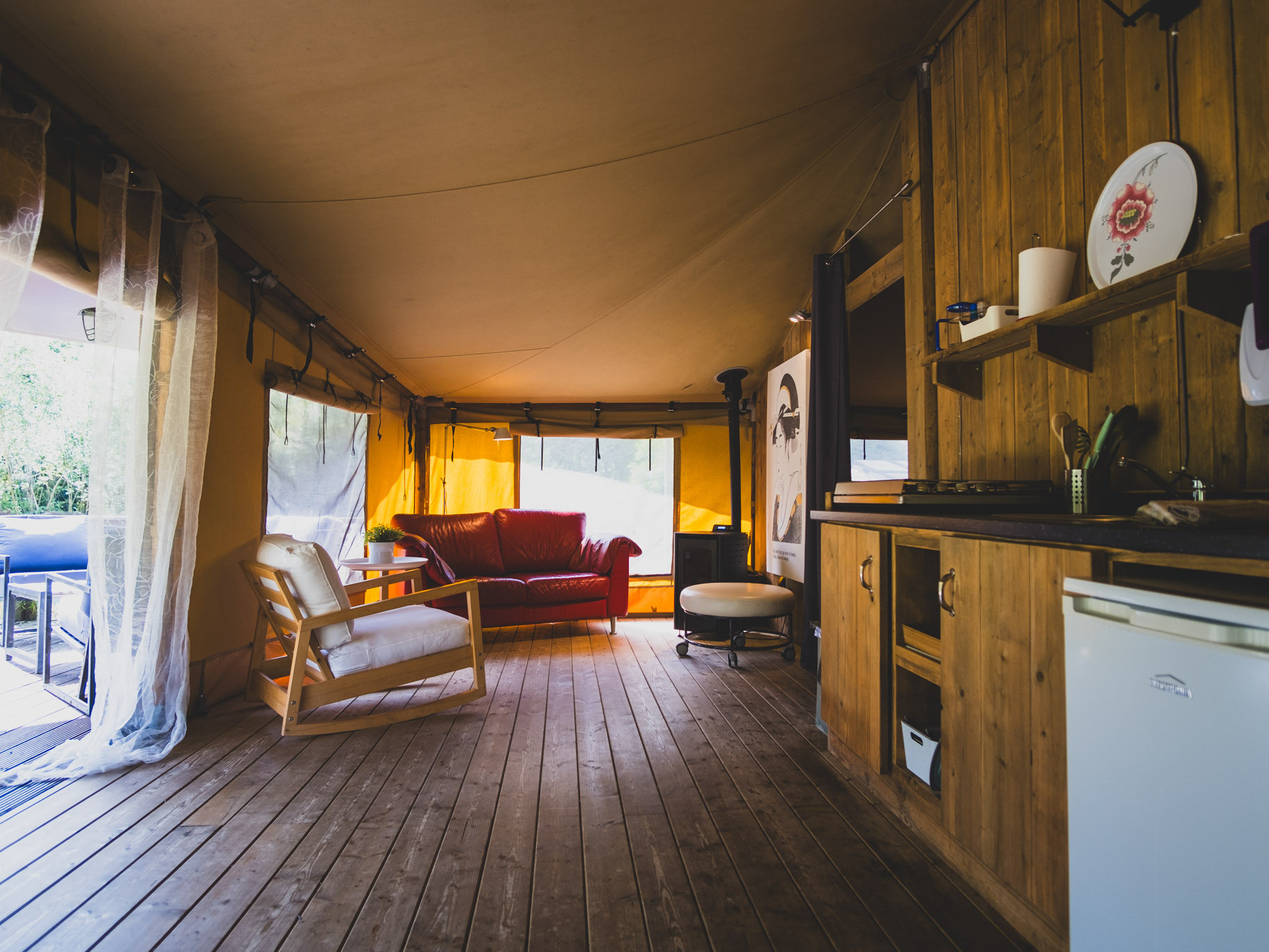 Interior of a tent with wooden floor, a red couch, a white armchair, and a small kitchen area.