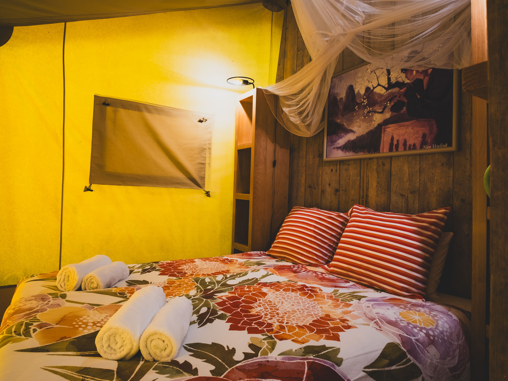 Bed with floral bedspread, striped pillows, rolled towels, wooden wall, and yellow fabric walls with window.