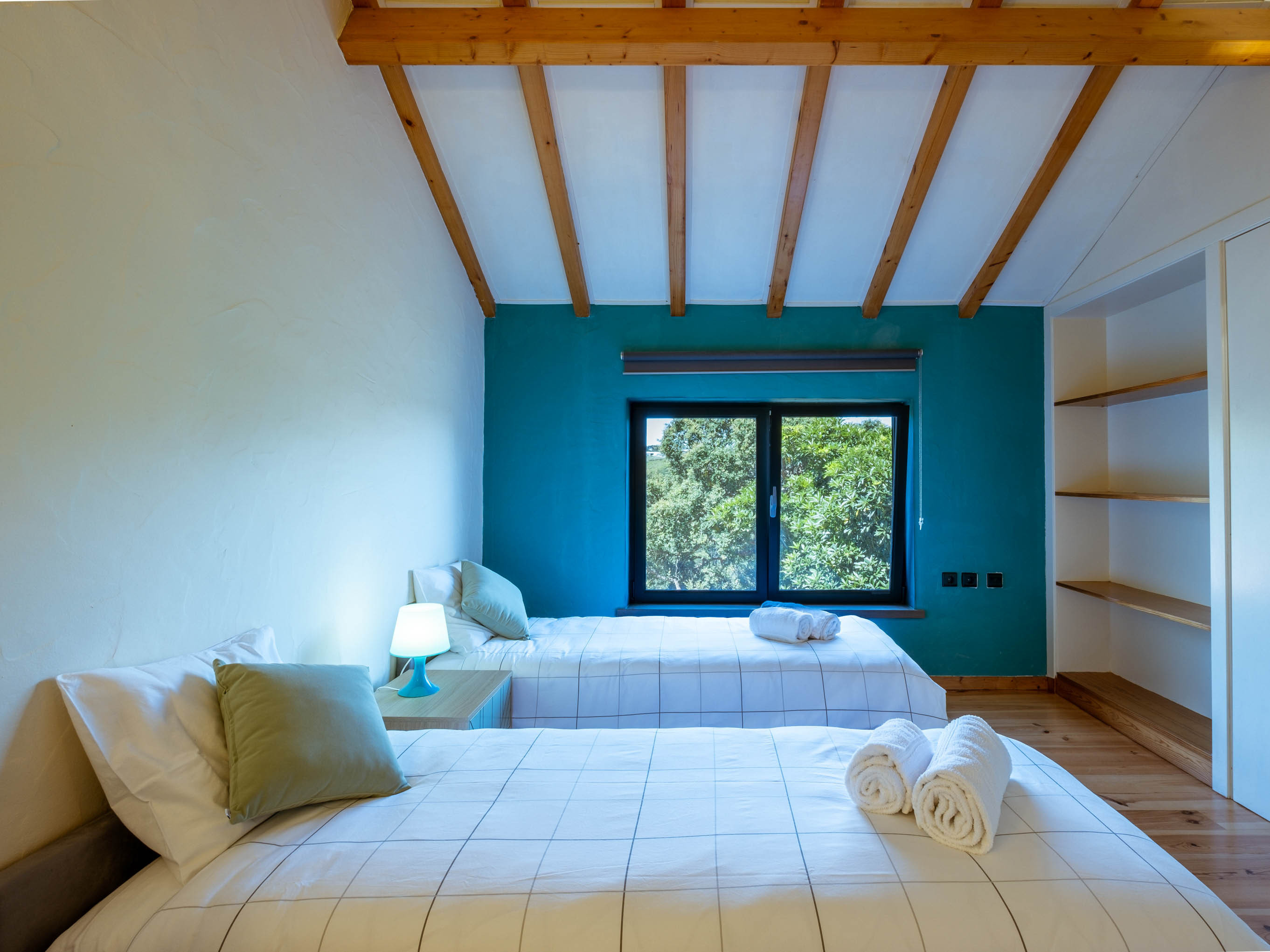 Bedroom with two single beds, teal accent wall, wooden ceiling beams, and a window showing trees outside.