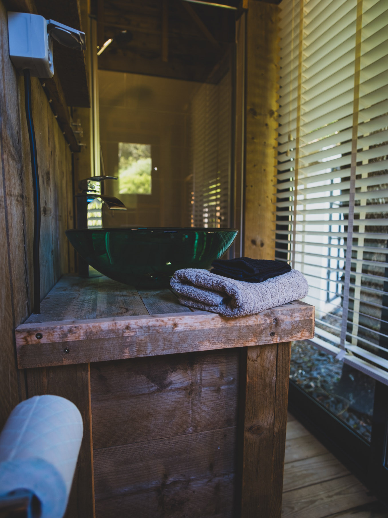 Rustic wooden countertop with green glass sink, stacked towels, window blinds, and partial bathroom interior.