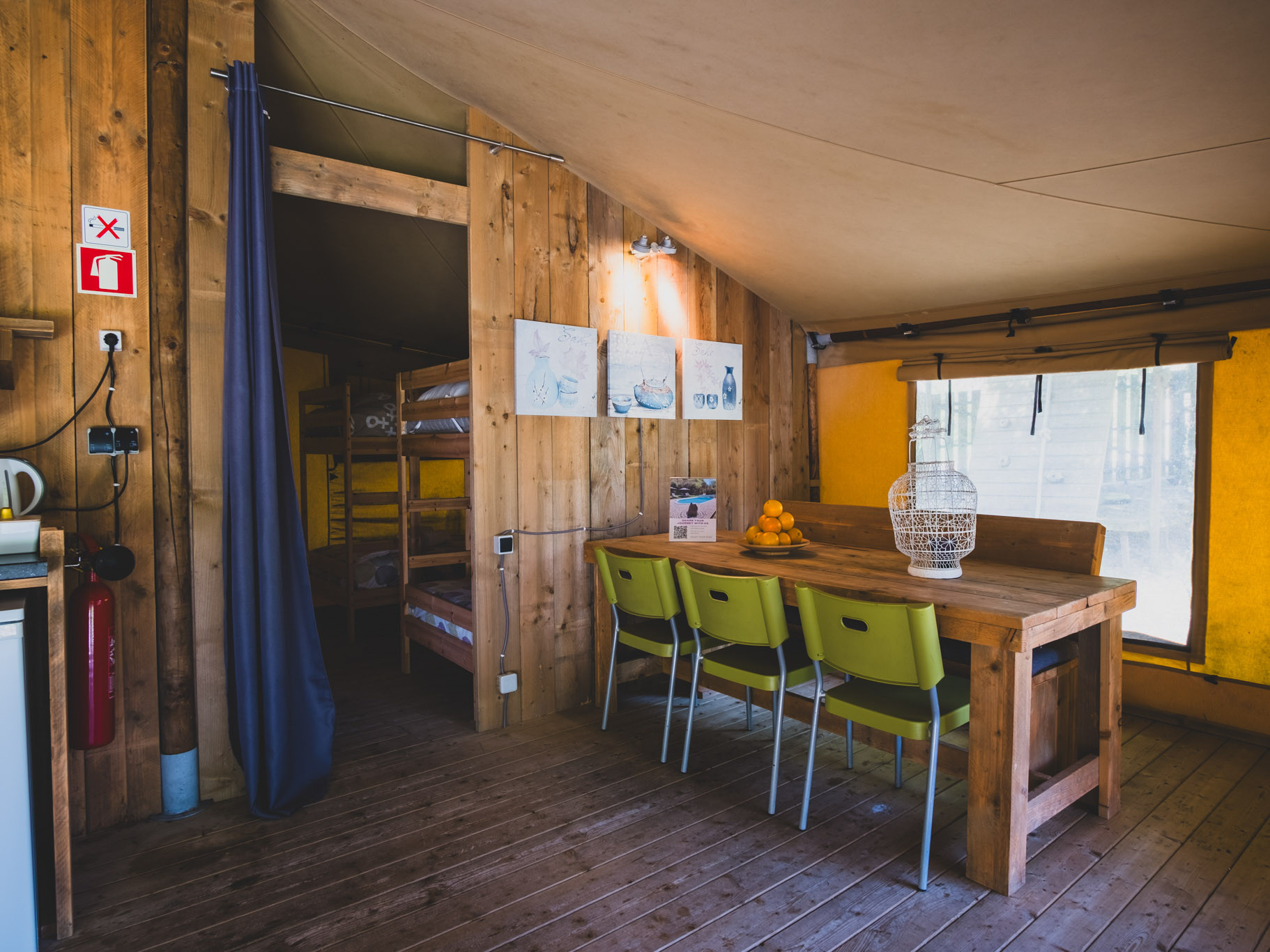 Wood-paneled dining area with wooden table, four green chairs, fruit bowl, and bunk beds behind curtain.