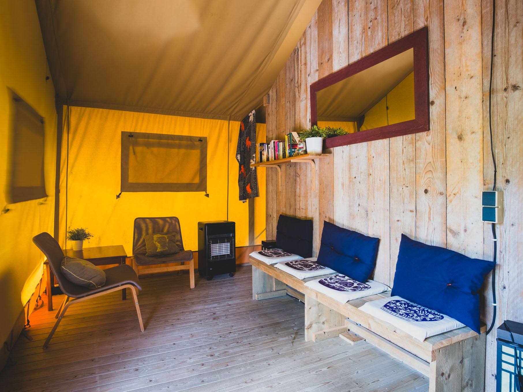Interior of a wooden tent with chairs, a bench with cushions, a heater, and books on a shelf.