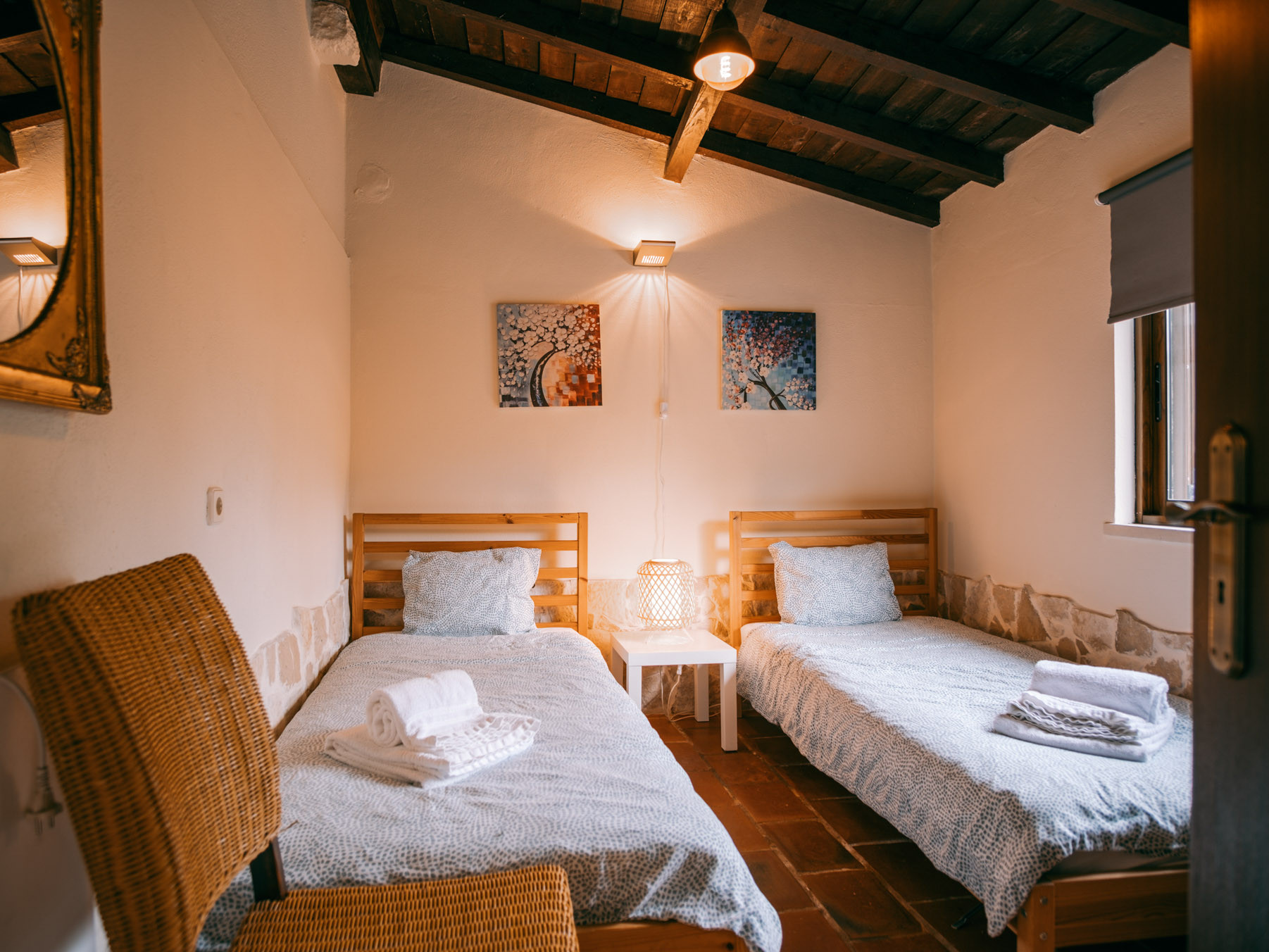 Room with two single beds, patterned bedspreads, pillows, two folded towels, a central side table, and wall art.
