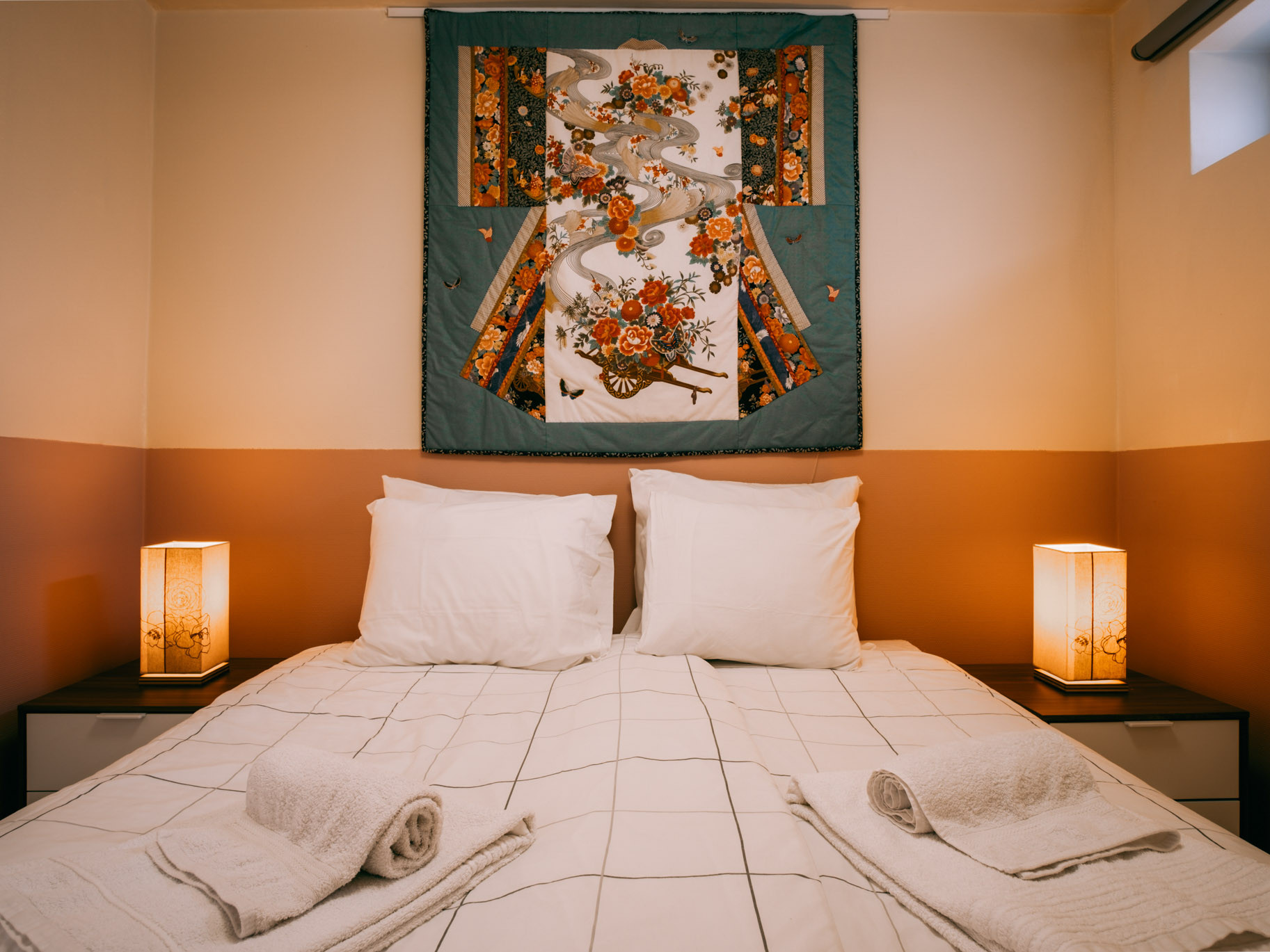 Double bed with white pillows and checked duvet, folded towels, bedside lamps, decorative textile hanging above.
