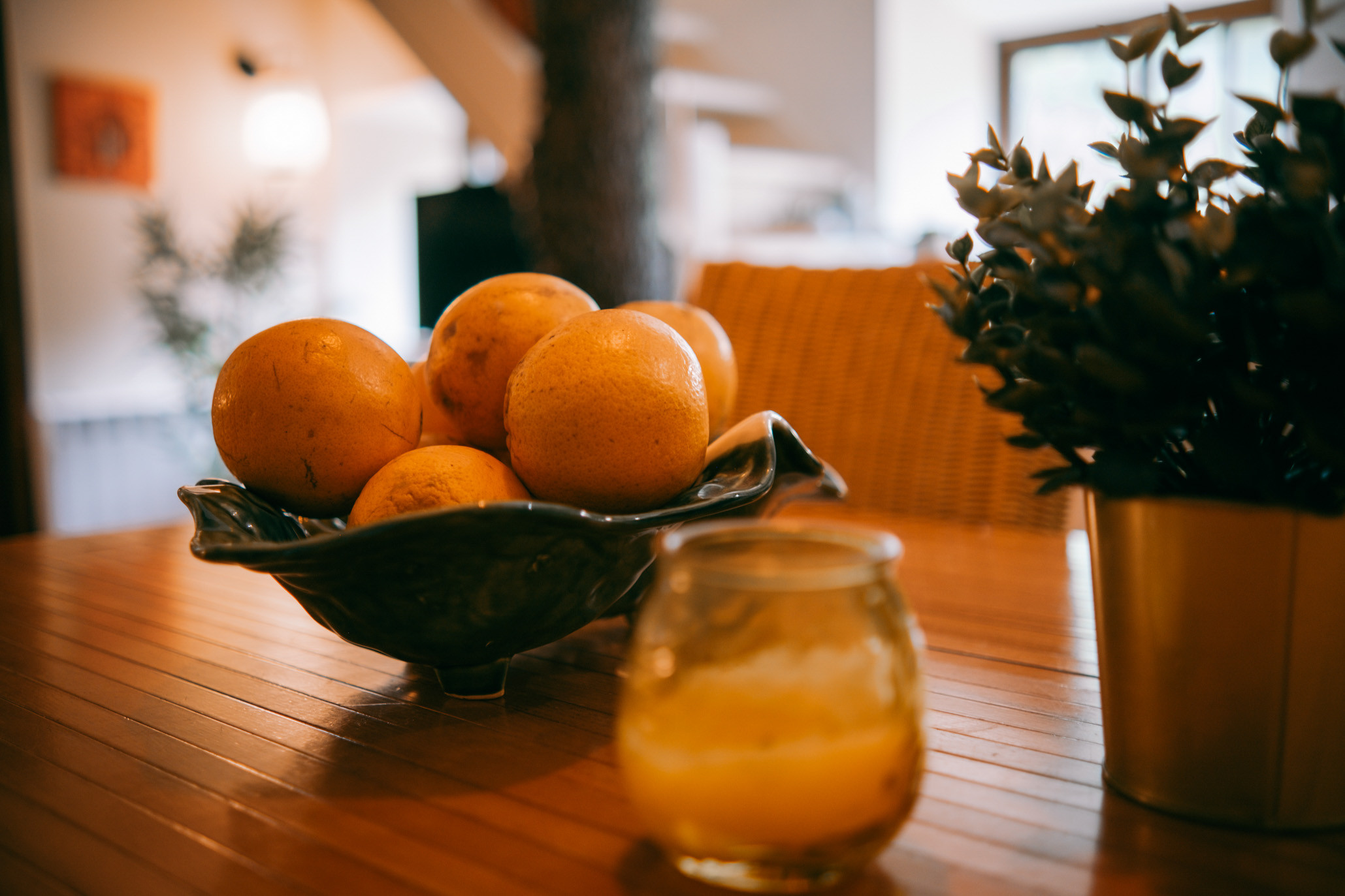 Bowl of yellow fruits on a wooden table alongside a small potted plant and a glass with juice.