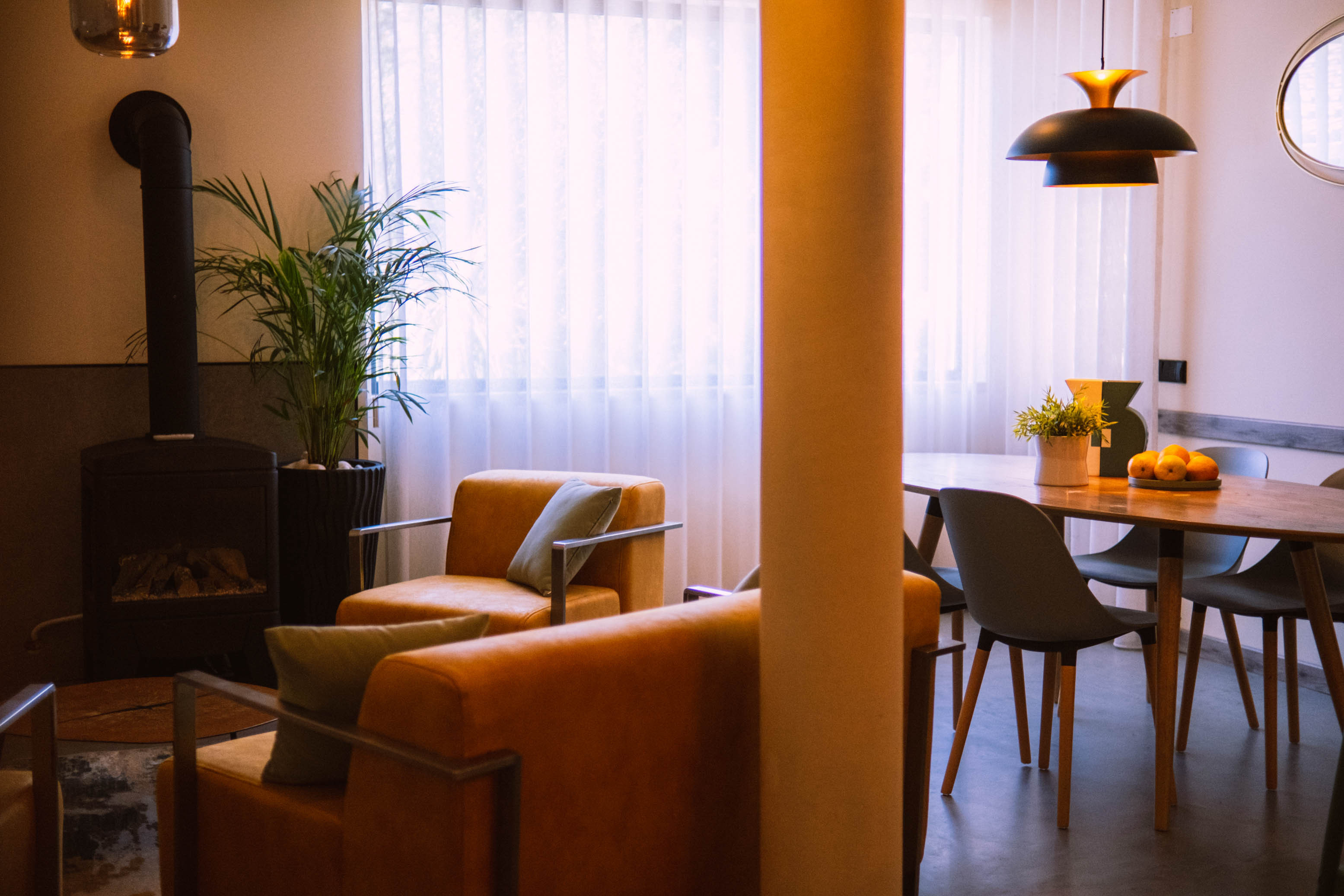 Cozy living area with orange armchairs, wood stove, potted plant, and dining table with chairs and fruit bowl.