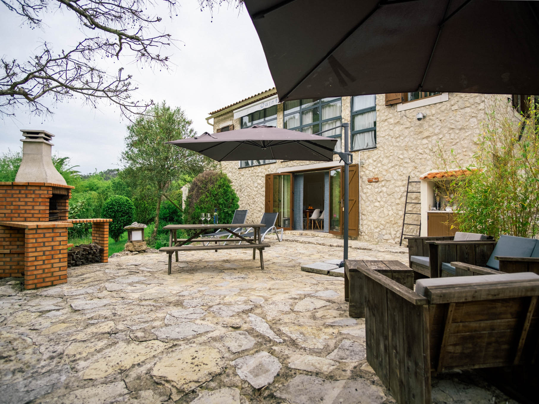 Stone patio with wooden seating, picnic table, umbrellas, and brick barbecue beside stone building.