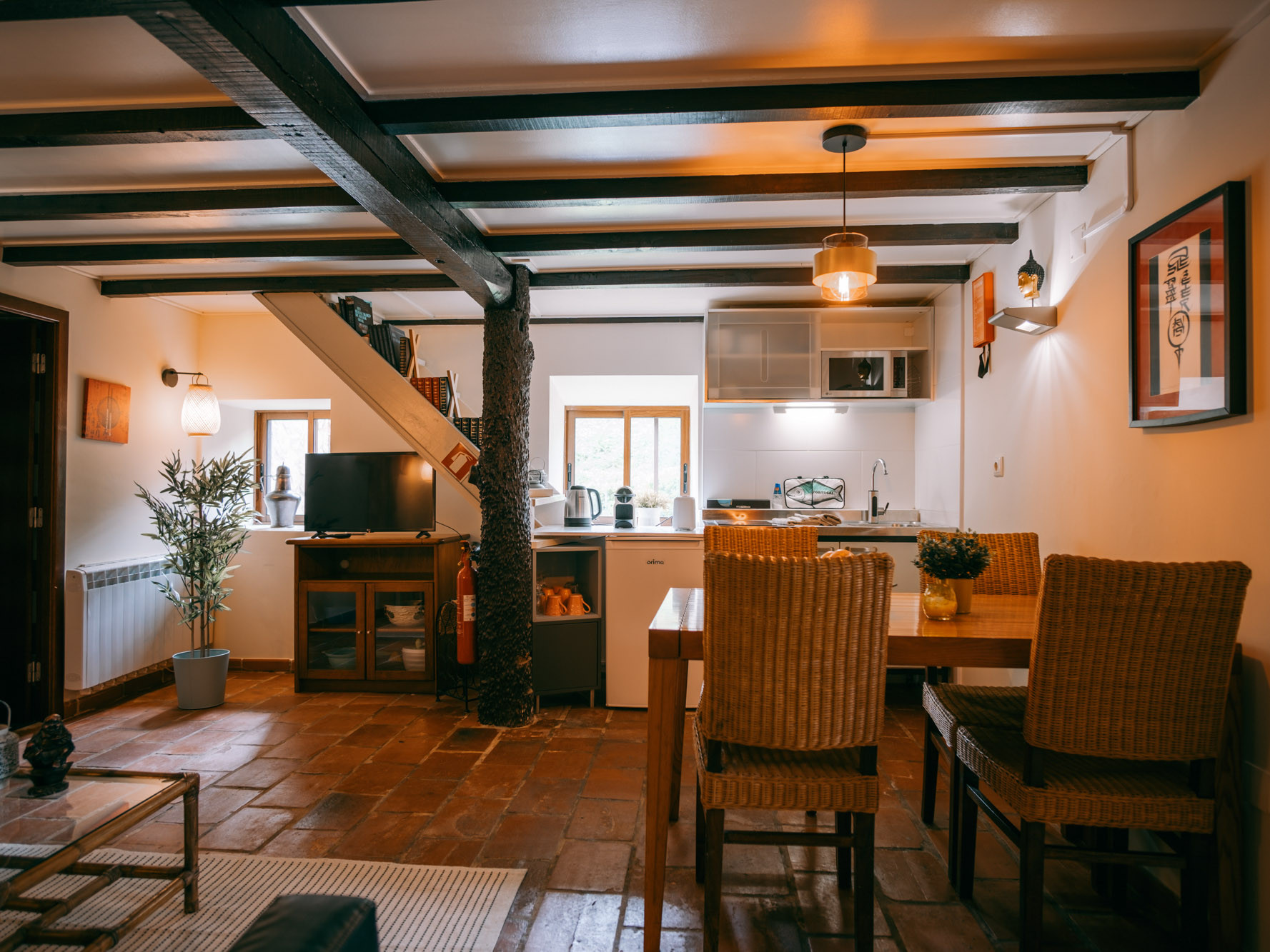 Cozy living space with dining table, wicker chairs, kitchen area, TV, and wooden ceiling beams.