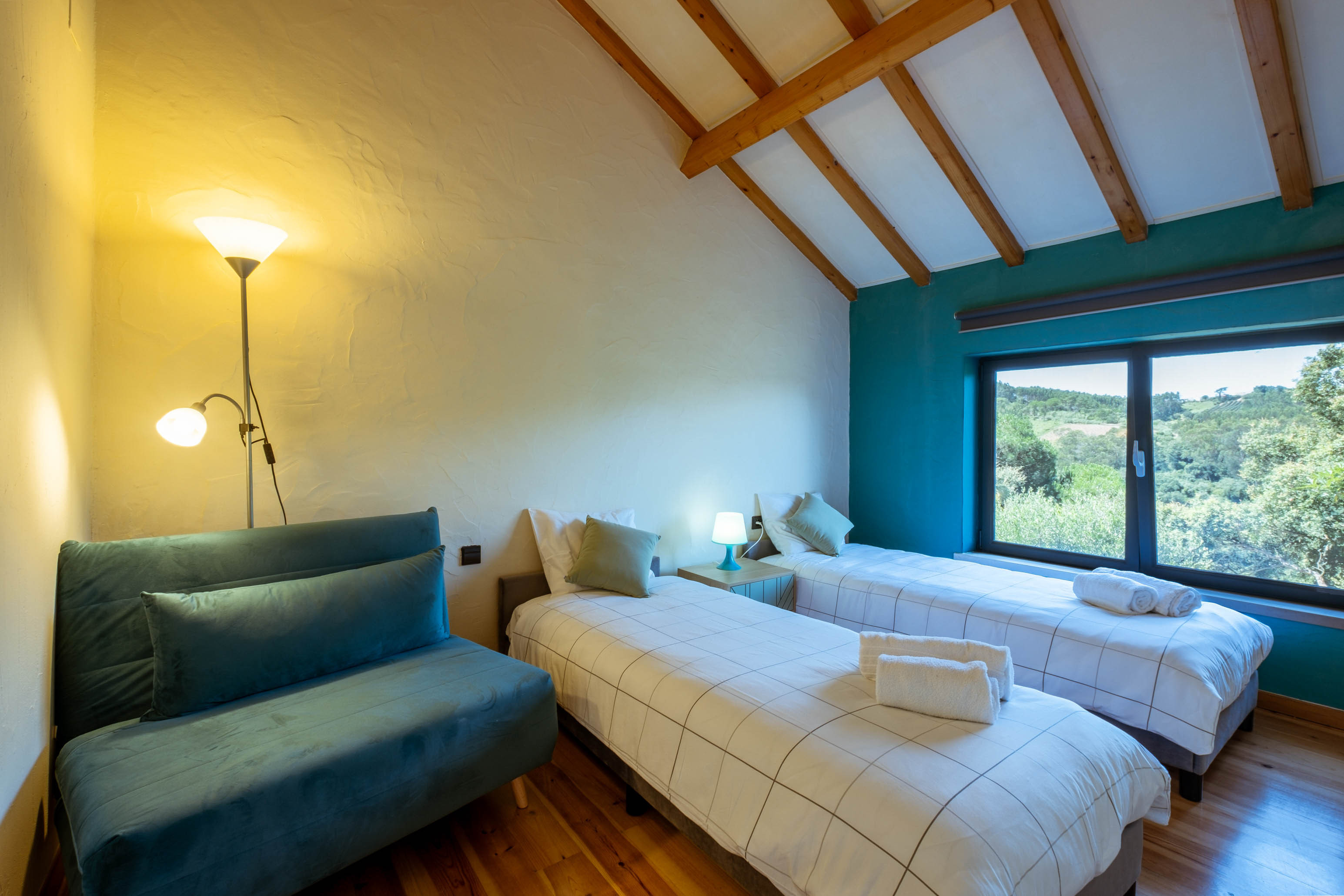 Room with two single beds, a blue sofa, wooden floor, and a large window showing green trees outside.