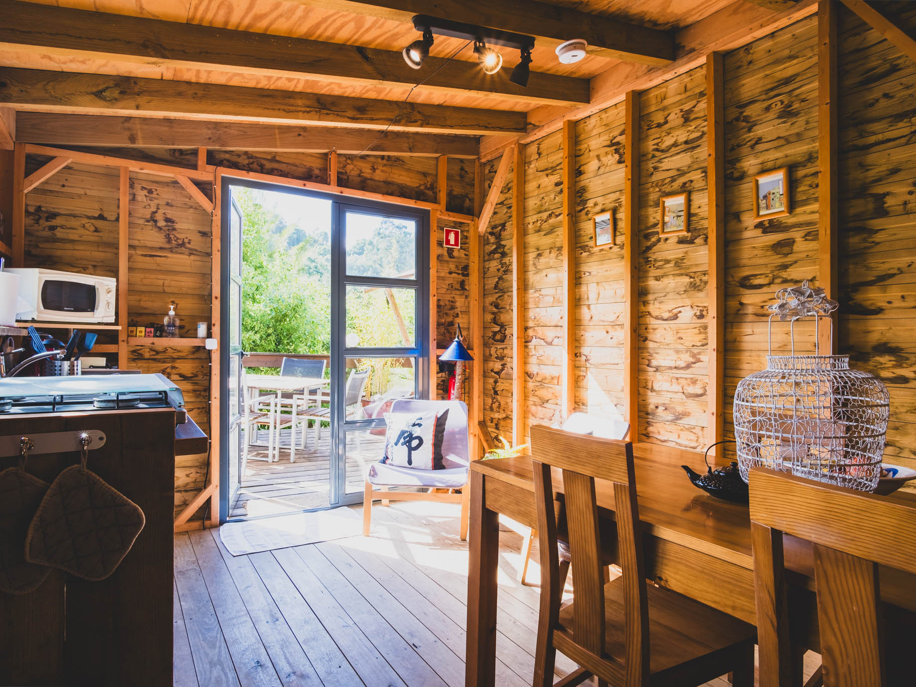 Interior of a wooden cabin with kitchen area, dining table, chairs, and open door to outdoor patio.