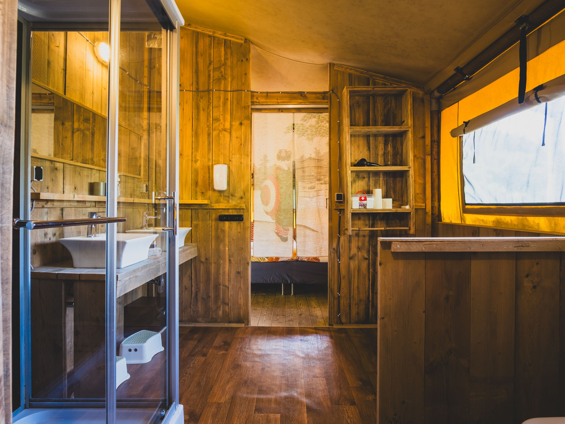 Wood-paneled interior with a bathroom sink, glass shower door, shelves, window with yellow curtain, and a distant bed.