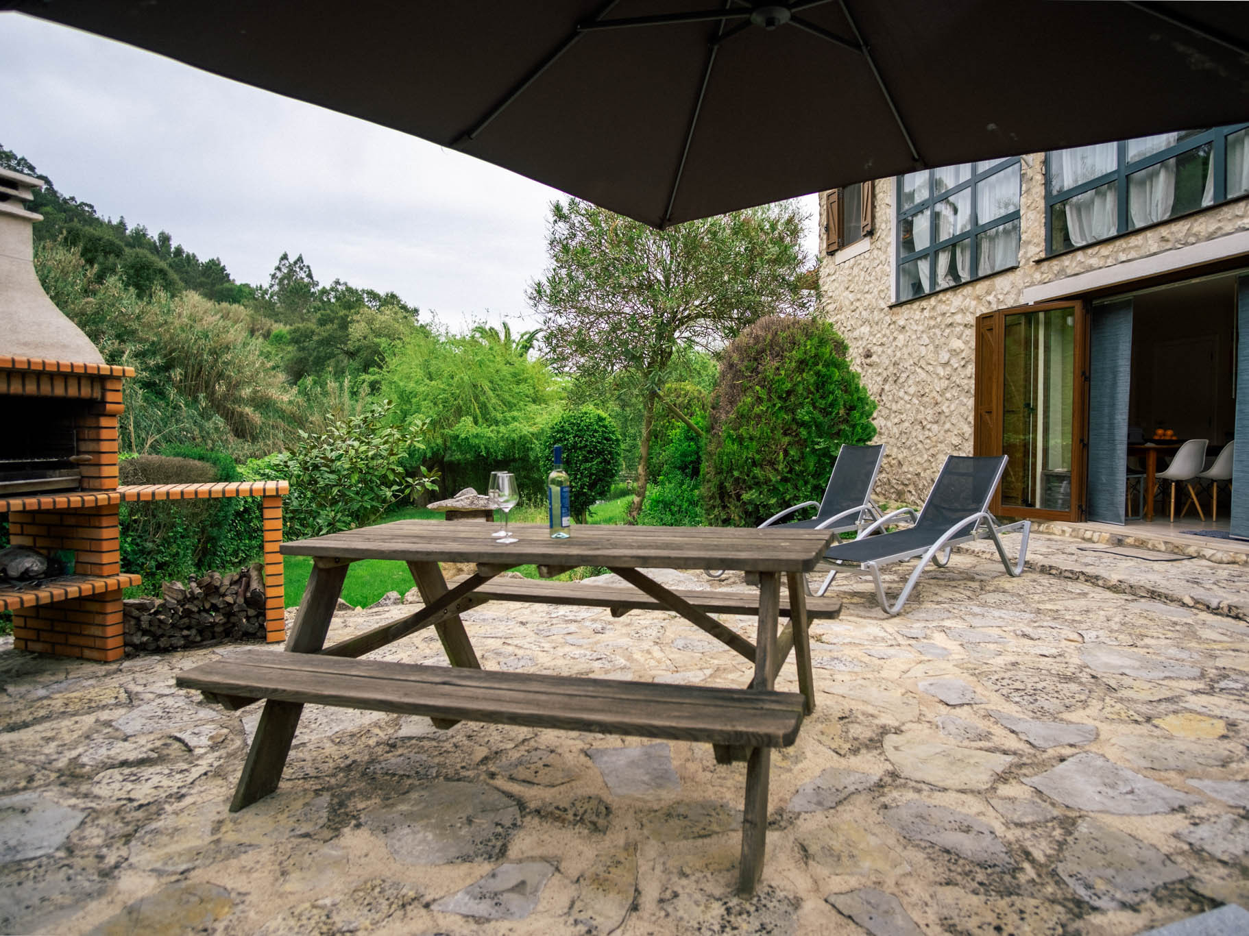 Stone patio with wooden picnic table, wine bottle and glasses, two lounge chairs, brick barbecue and garden.
