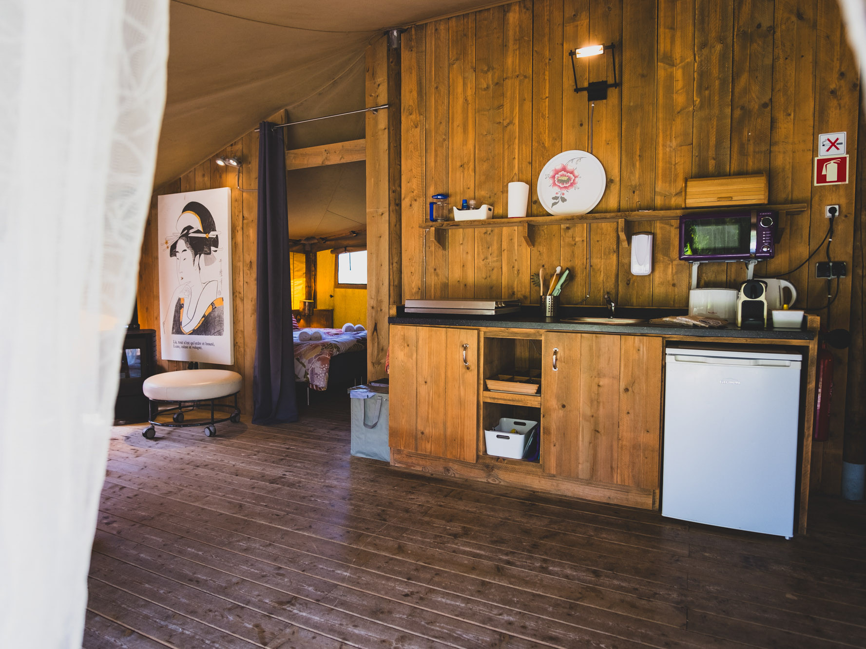Wood-paneled kitchenette with small fridge, microwave, sink, and utensils in a rustic interior space.
