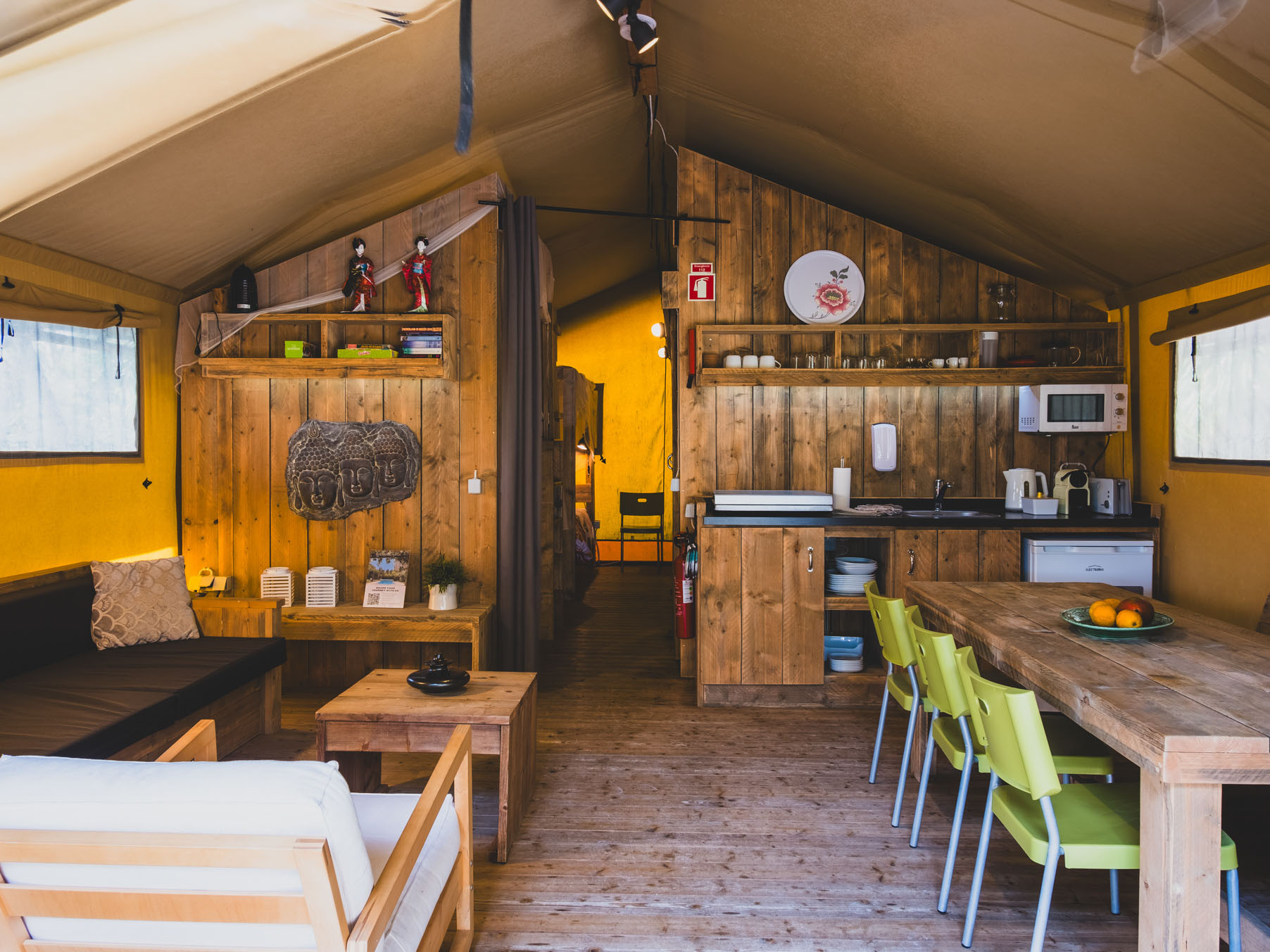 Interior of a tent with wooden furniture, kitchenette, dining table with green chairs, and decorative items.