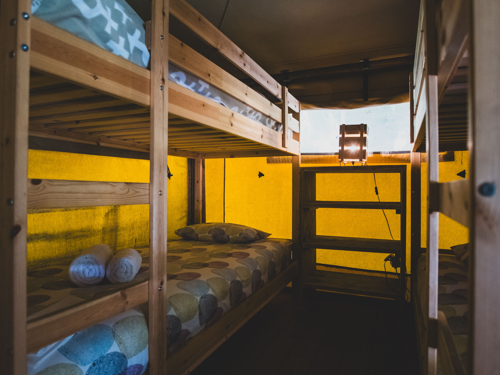 Wooden bunk beds with patterned bedding inside a yellow tent structure, towels on lower bed.