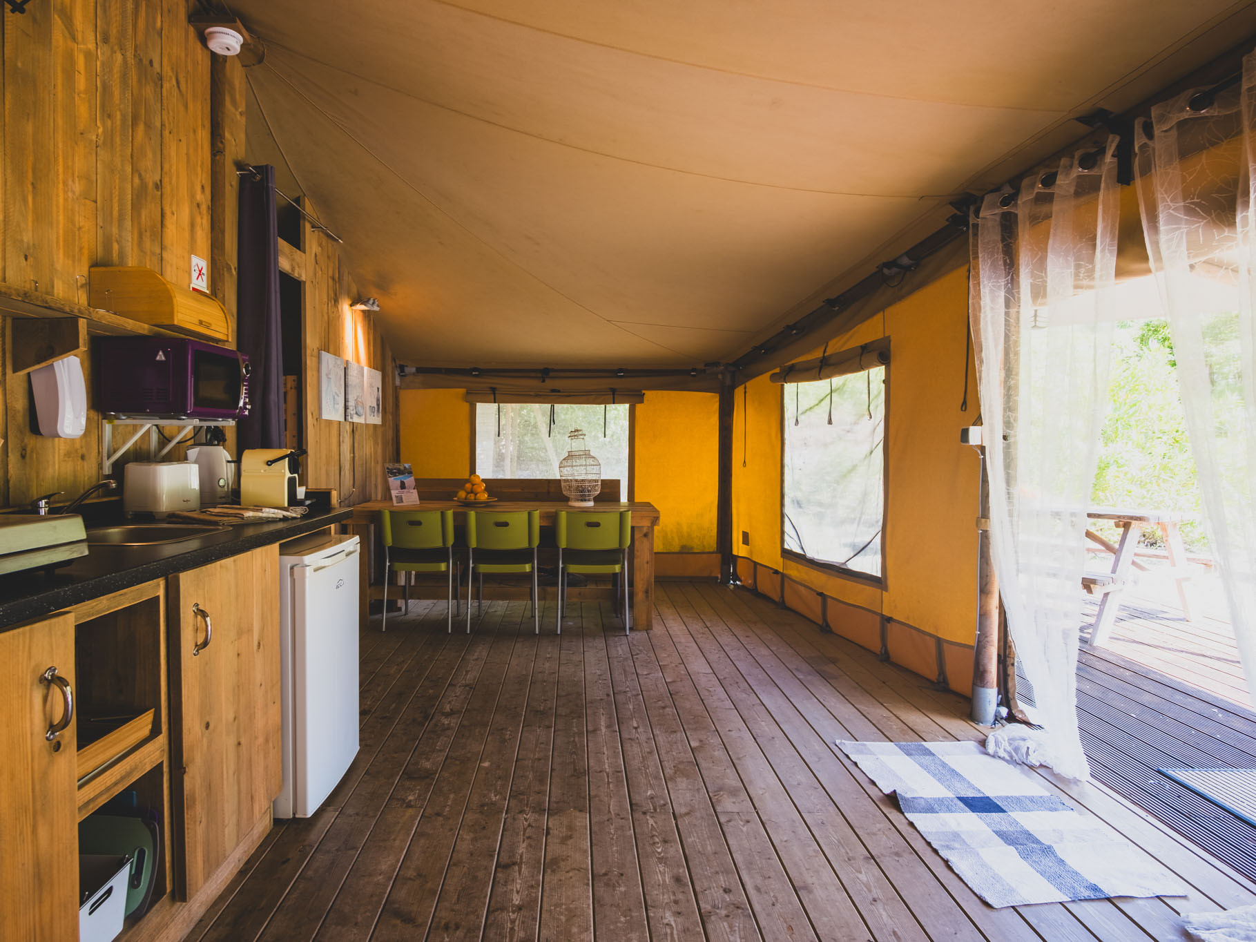 Interior of wooden cabin with kitchenette, dining table, green chairs, and large windows with sheer curtains.
