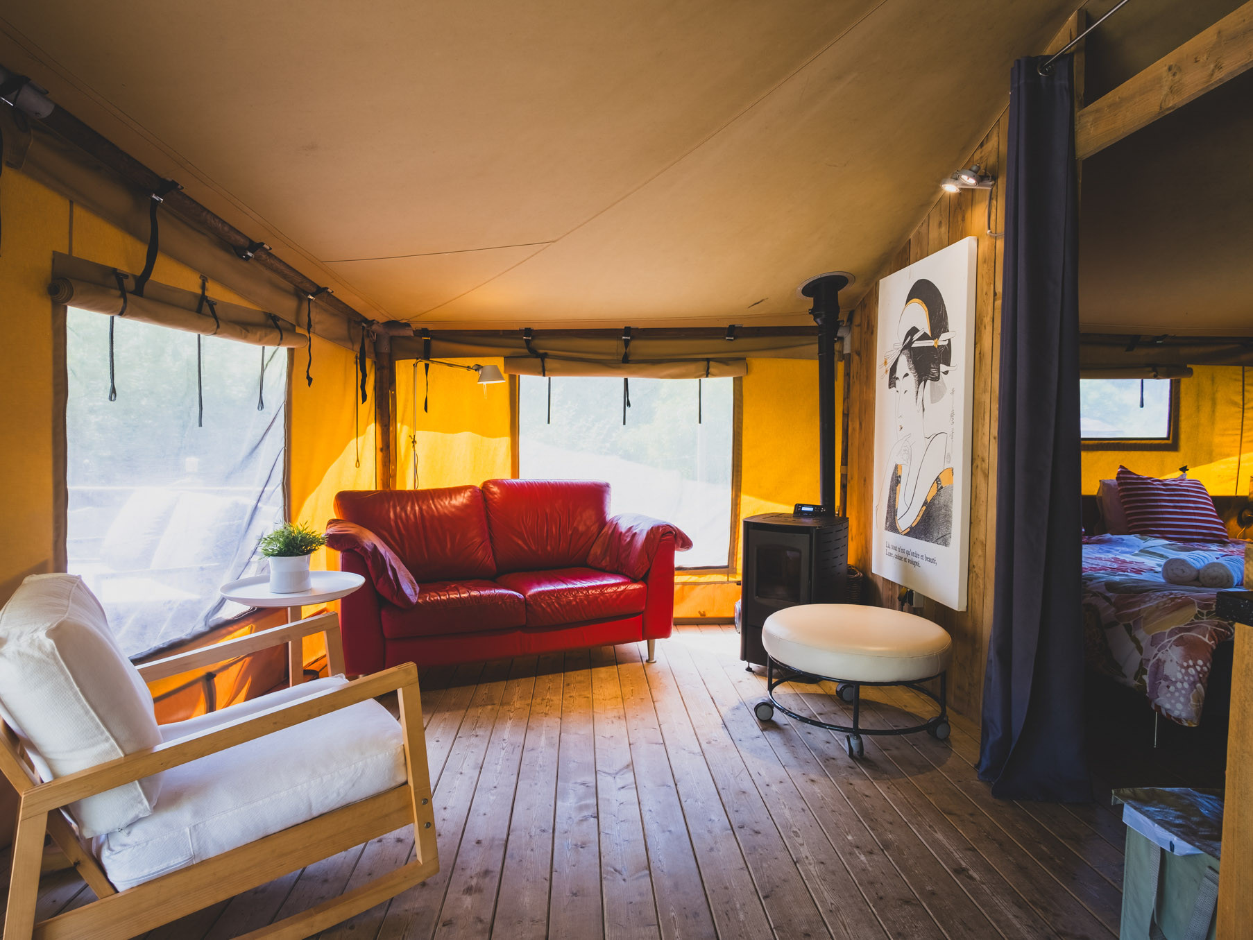Interior of a cozy tent with wooden floor, red sofa, armchair, small table, and a bed