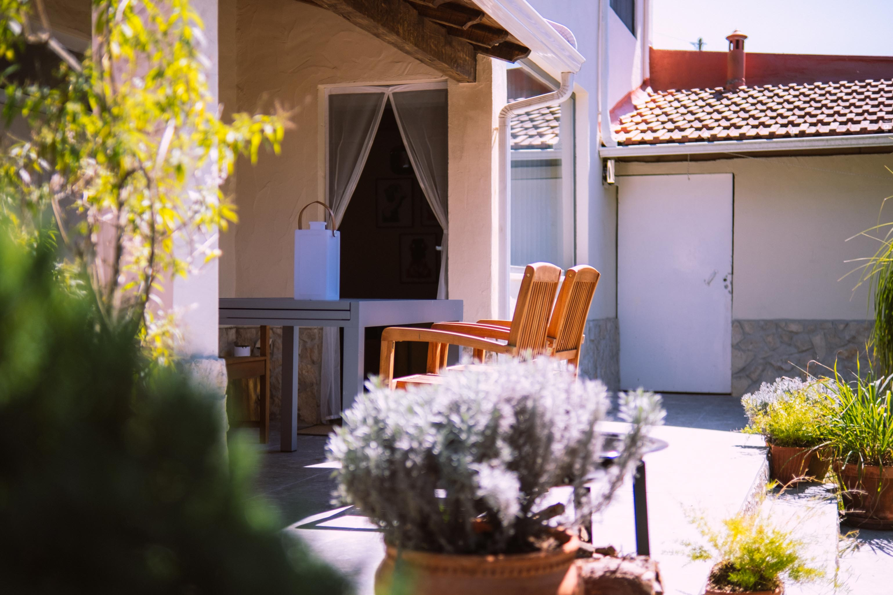 Patio with wooden chairs, table, and potted plants outside a building with tiled roof.