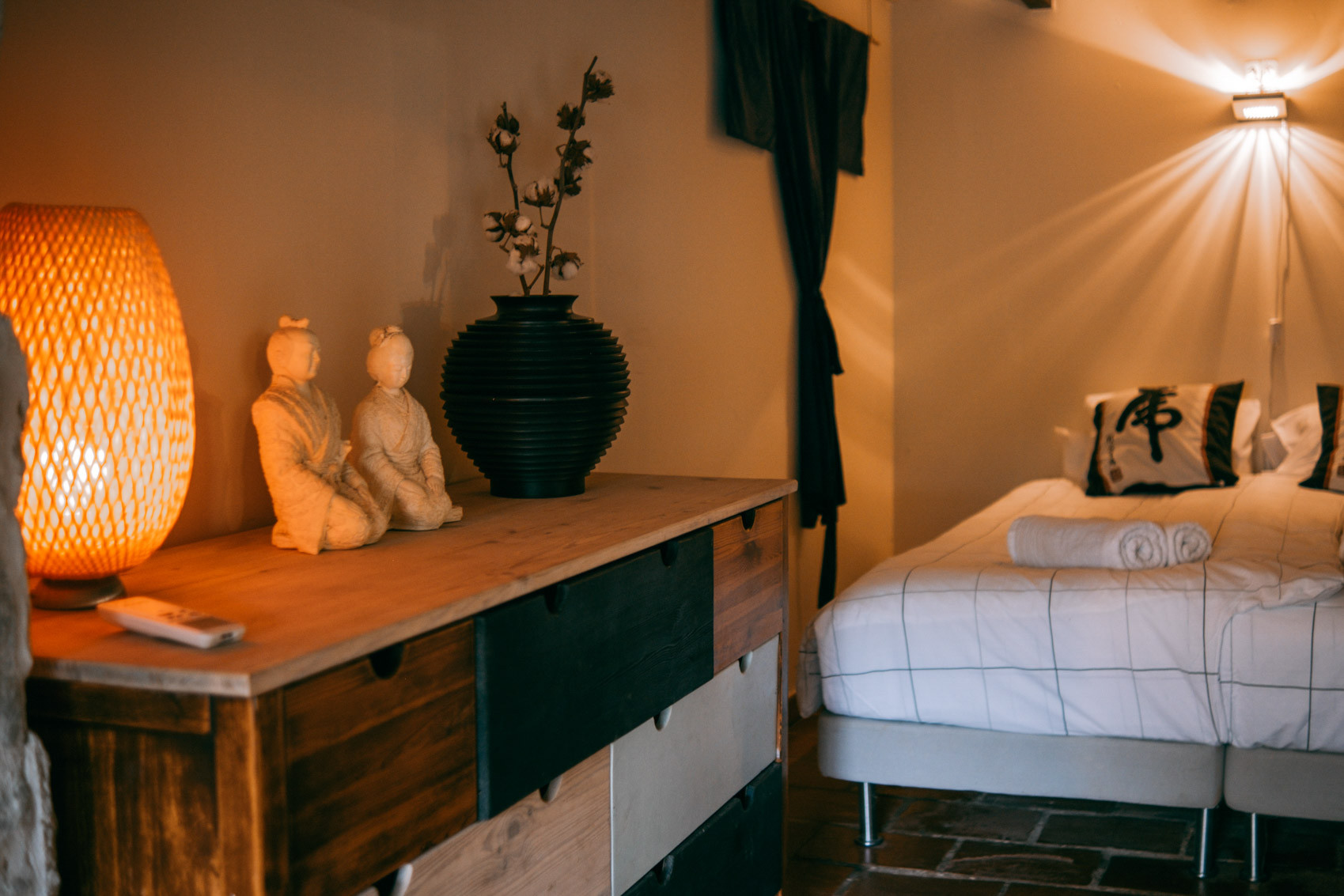 Bedroom corner with twin beds, rolled towels, dark curtains, wooden dresser with sculptures and vase, warm lamp light.