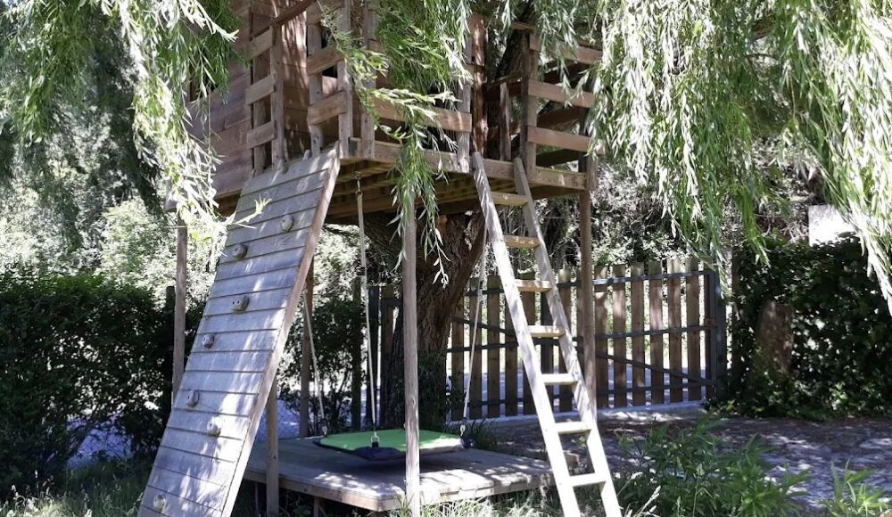 Wooden treehouse with climbing wall, ladder, and swings under leafy willow branches