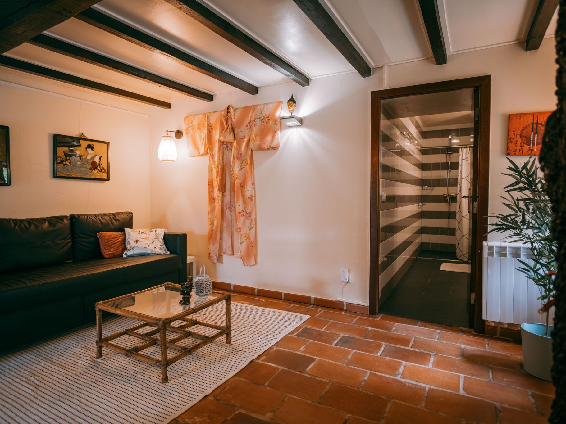 Living area with black sofa, glass coffee table, wall art, hanging garment, and view into striped tiled bathroom.